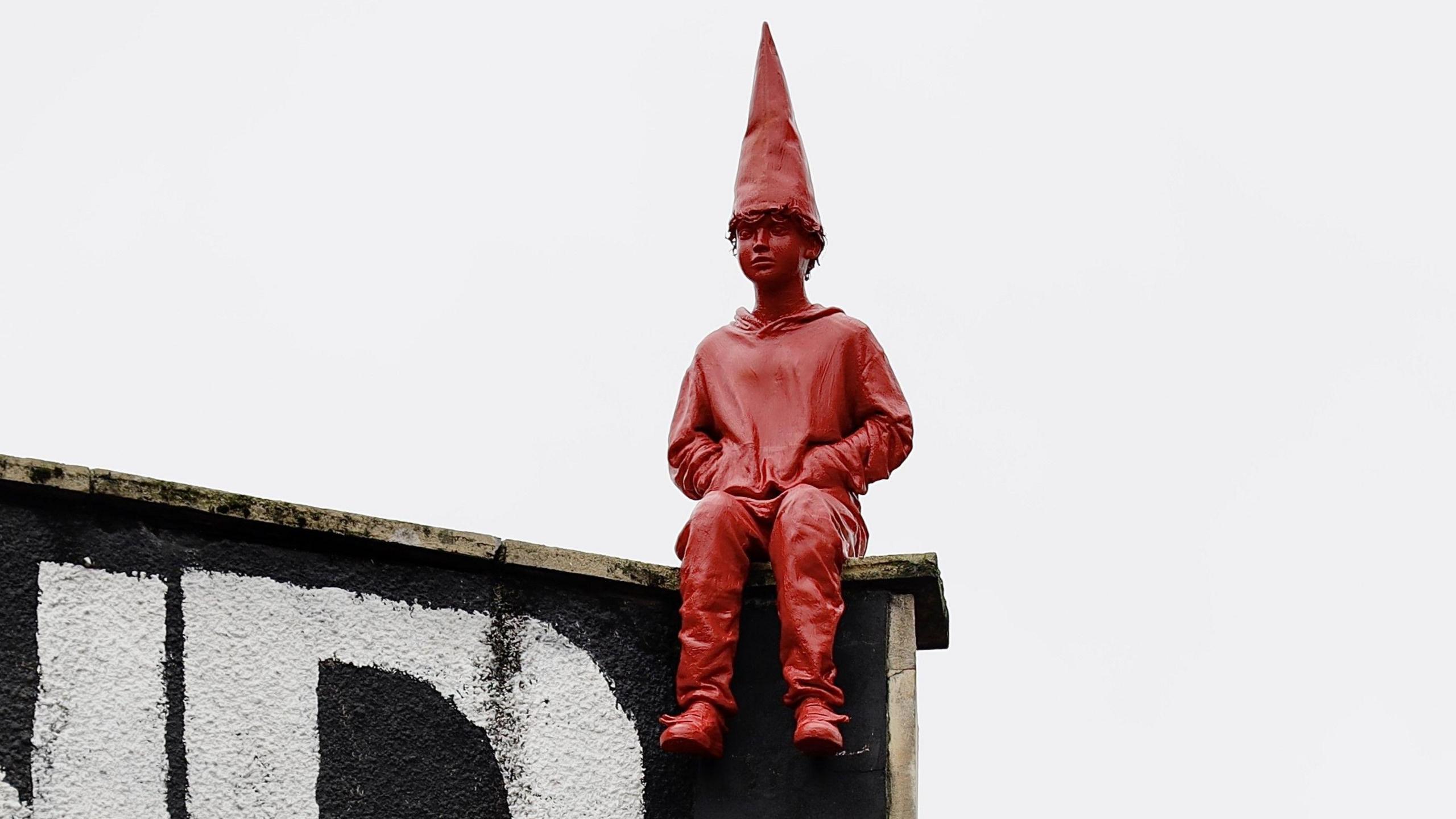 A close up picture of the "dunce" statue. It is entirely red, and shows a boy with curly hair, wearing a red dunce hat and a hoodie, sat on the corner of the building. 