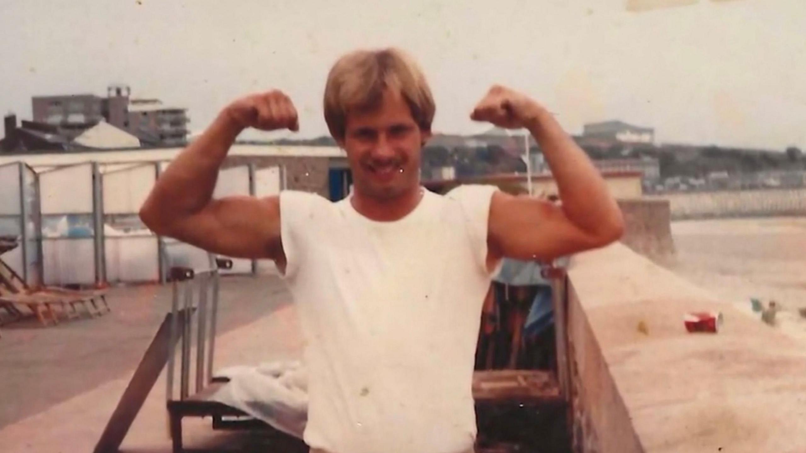 Danny Bowers, in an old photograph taken with colour film. He has both his arms raised, flexing his muscles, and is wearing a white t-shirt.