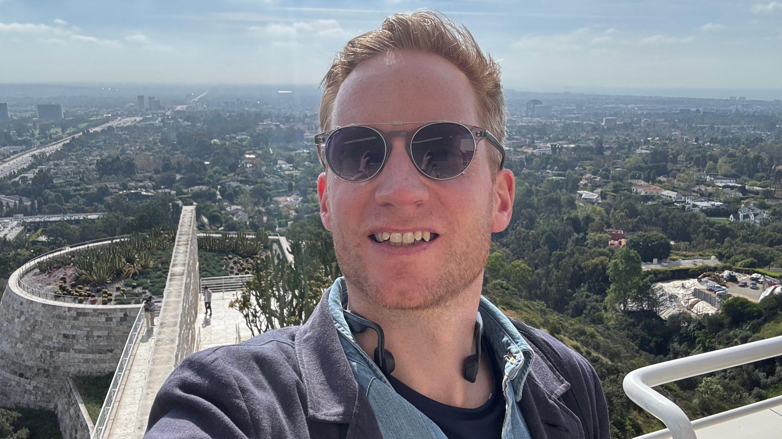 Kieran wearing sunglasses taking a selfie from an elevated outdoor terrace, with a sprawling cityscape and green hills in the background. A curved stone walkway and landscaped rooftop garden are visible below under a bright, hazy sky.