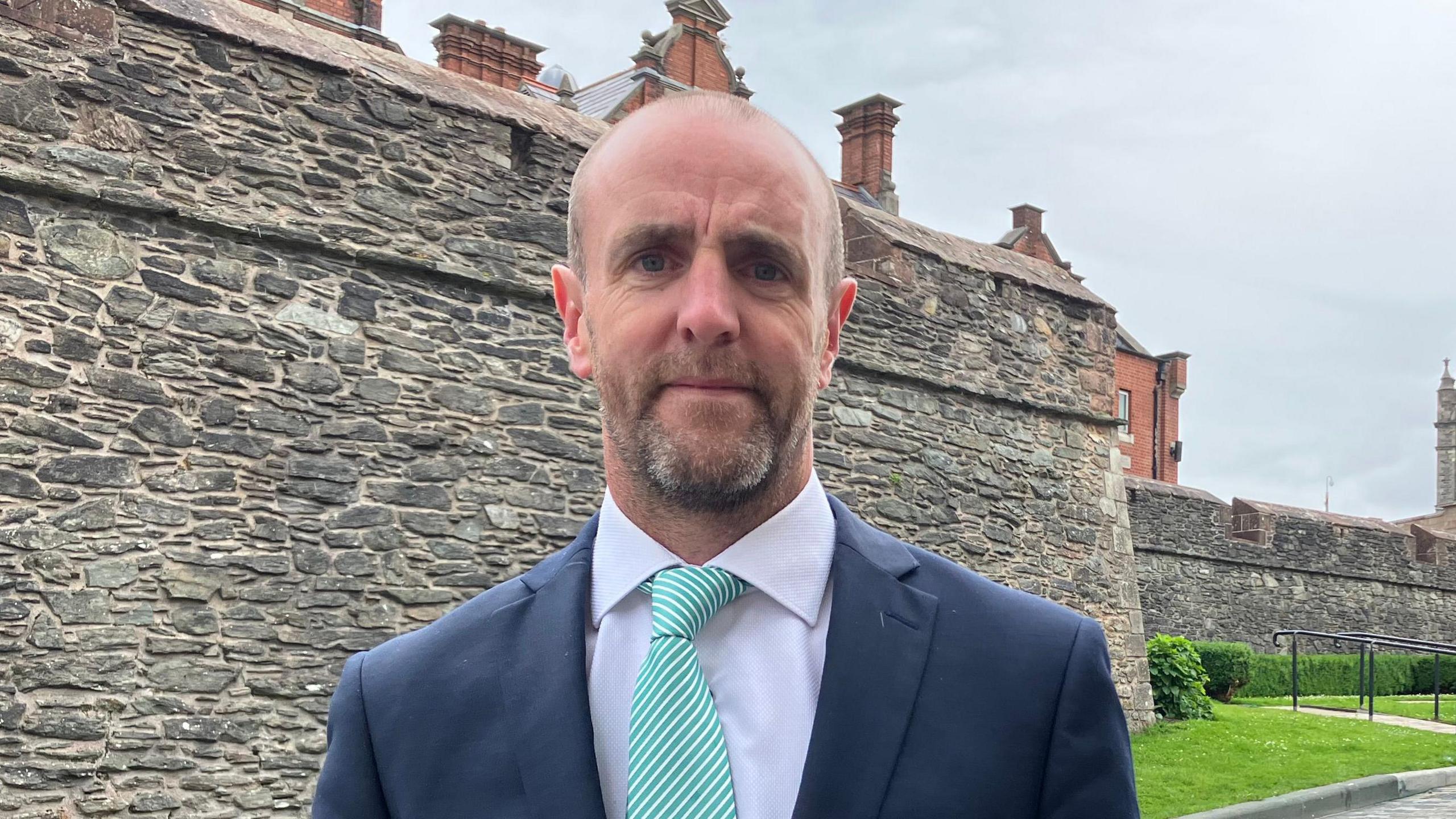 Mark H Durkan is wearing a green and white striped tie, white shirt, and a navy blazer. He is standing near to Derry's historic walls.