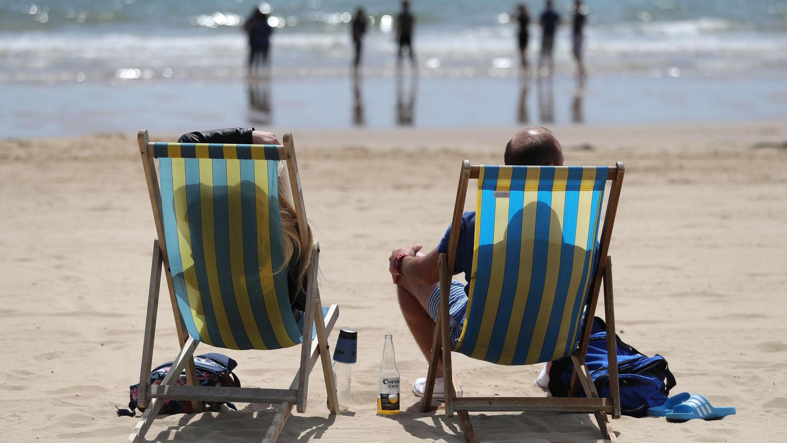 A rear view of a man and woman sitting in two striped deckchairs on a sandy beach. They are looking towards the sparkling sea where there are seven people standing in the shallow water.