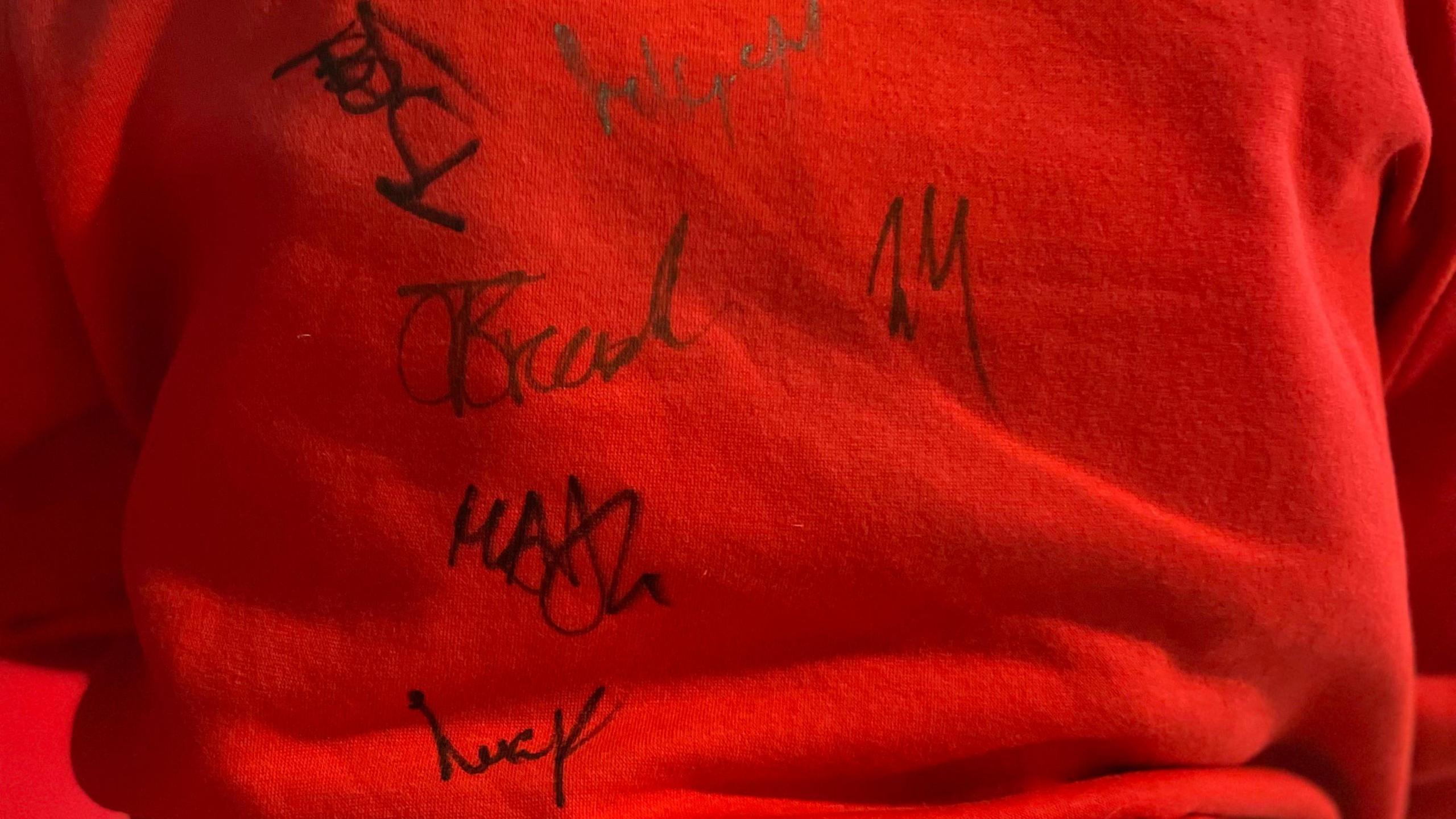 A person is wearing a red sweatshirt covered with multiple signatures in black and silver ink across the back. The signatures are scattered over the upper and middle sections. The background is softly blurred, suggesting an indoor setting with natural light from a window. The focus is on the signed sweatshirt, likely a keepsake or memorabilia item