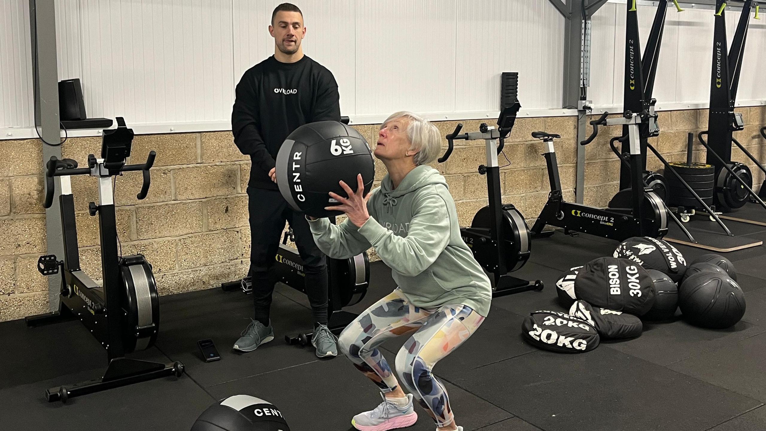 Celia Duff is in a gym and is squatting while holding a 6kg black ball. She has Pink and grey trainers, patterned pink, grey, yellow and blue gym leggings and a light blue hoodie with Ovrload written in the center. She has studded earrings and short grey hair with a side fringe. Watching her workout with his hands clasped in front of him is personal Trainer Mike Creamer. He wears black gym pants, a black sweatshirt with Overload  in the centre and has short dark hair and facial hair. 