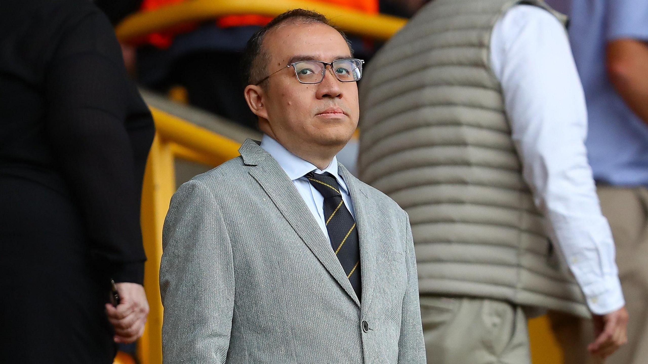 Jeff Shi at Molineux in a grey suit, a shirt and tie and dark-rimmed glasses, looking in the direction of the camera