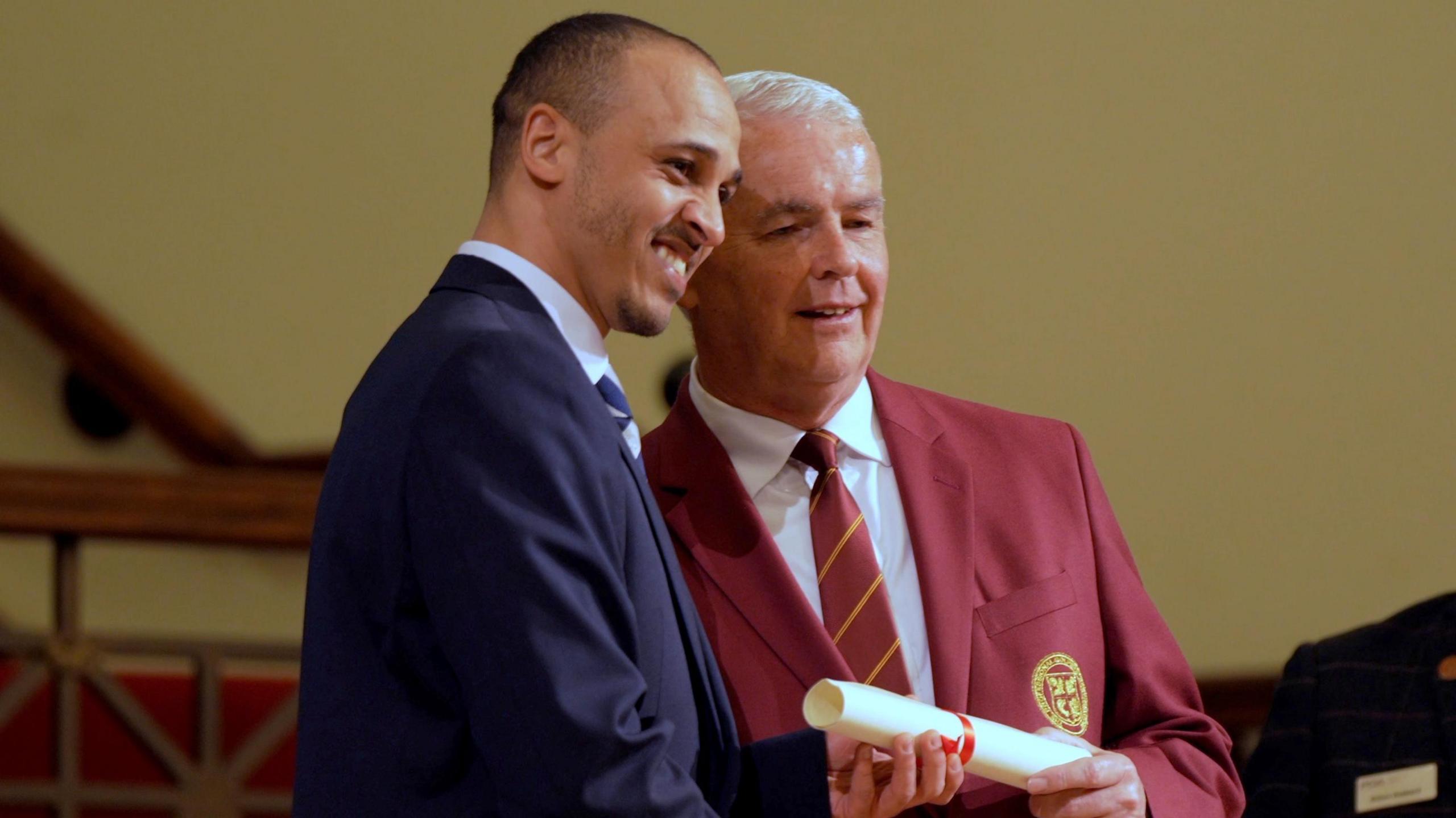 Peter Odemwingie wears a dark blue jacket and tie and smiles while receiving his degree scroll from a man wearing a maroon jacket and tie