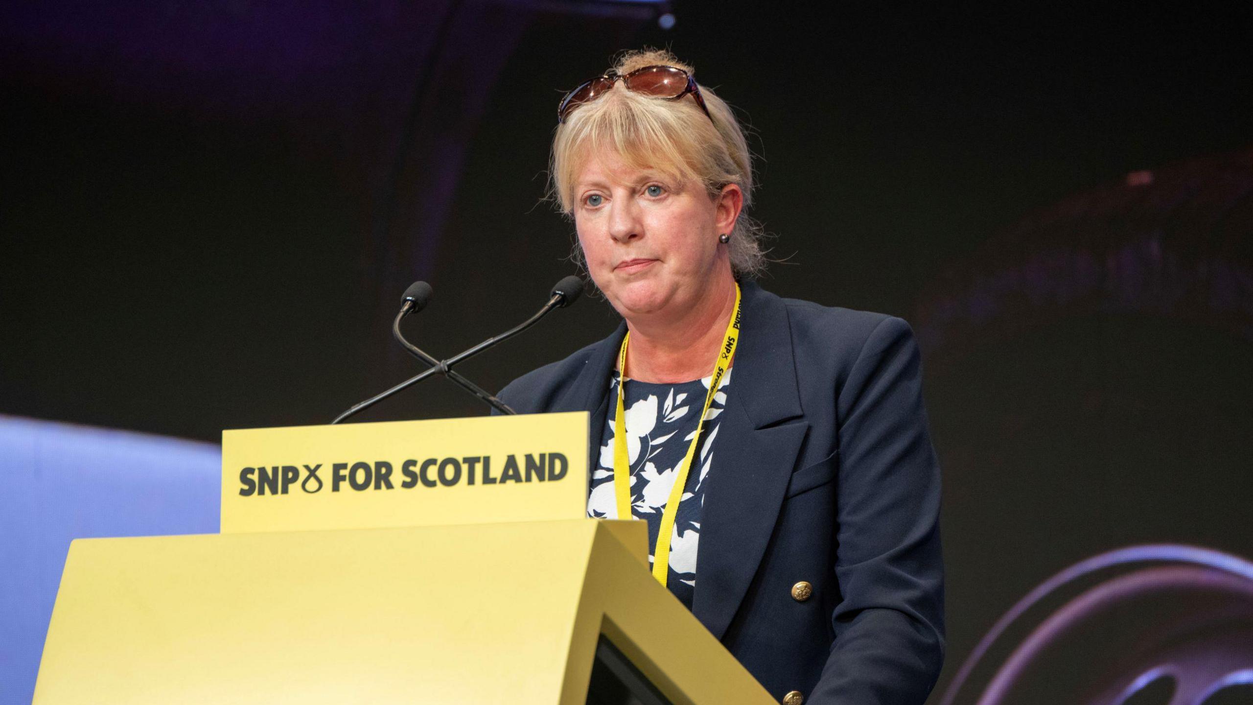 Shona Robison, with glasses resting on top of her head and dressed formally, making a speech at an SNP conference. A yellow conference lanyard is around her neck and the yellow podium she is standing behind says SNP FOR SCOTLAND on it 