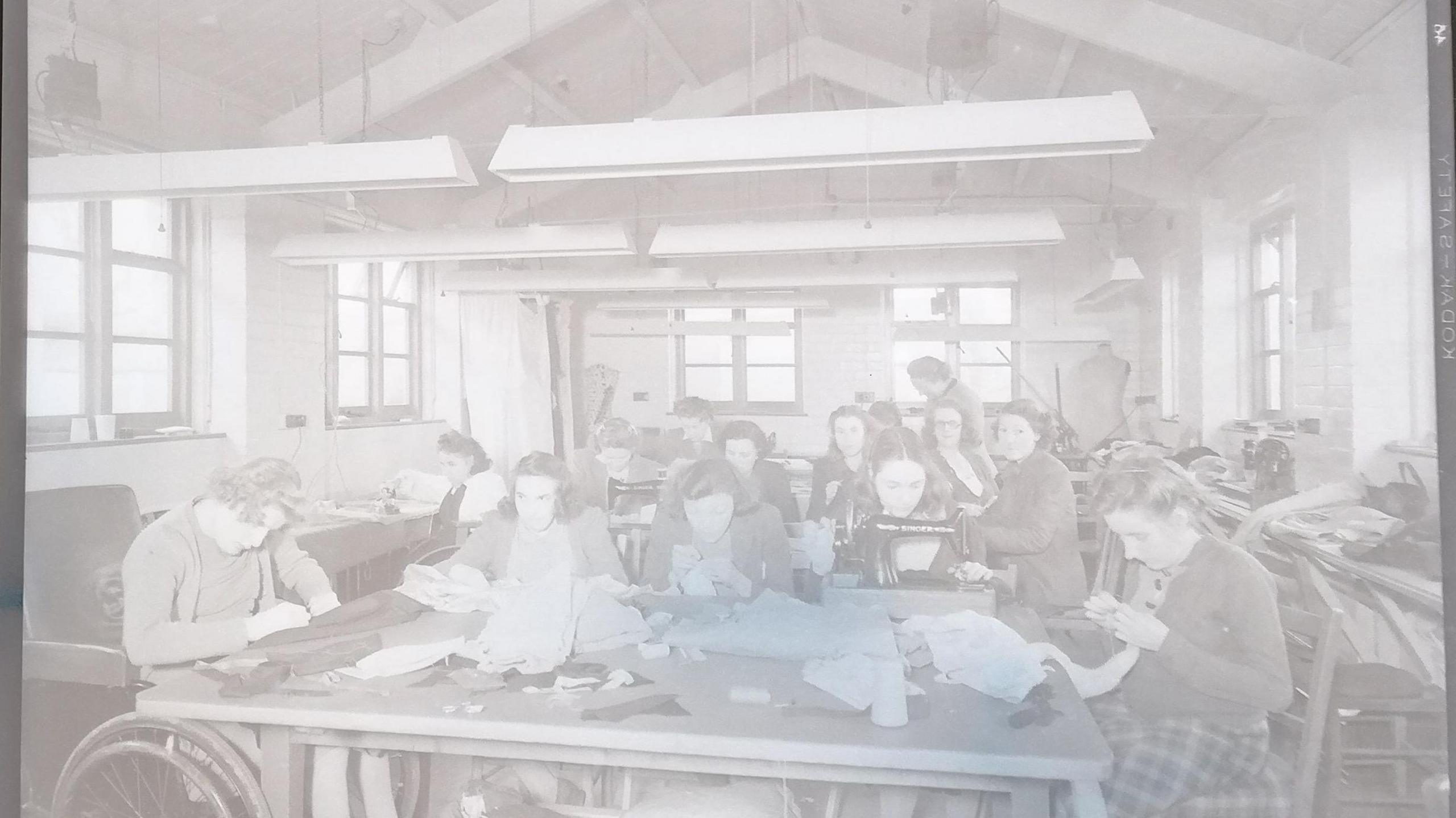 A negative shows residents of St Loye's College and School of Occupational Therapy, taking part in a sewing workshop. It is hard to distinguish everything in the image but women or girls are sewing by hand and one can be seen sitting in front of a sewing machine. To the left of the photo a wheelchair user is working on a dark piece of fabric.