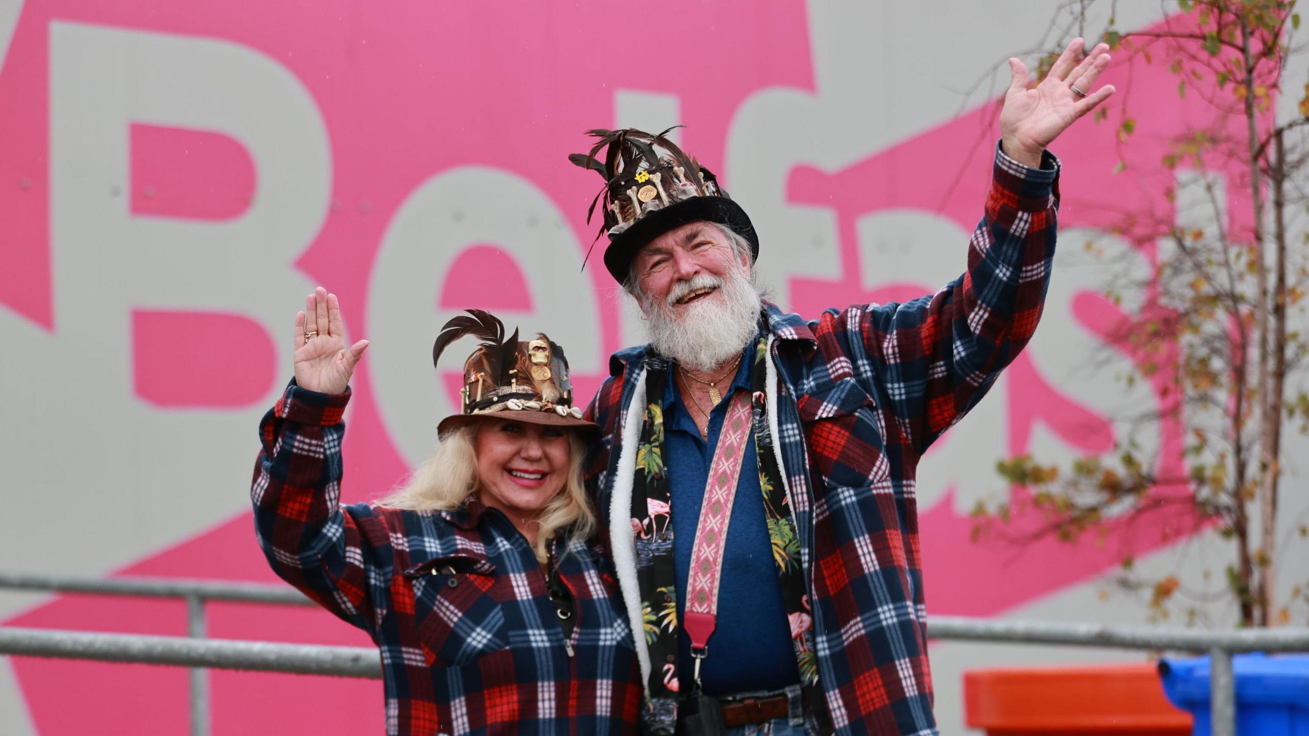 A man and woman in red and blue check shirts waves towards the camera. The man has white hair and a beard and is wearing a blue top and brown hat with feathers. He has his arm around a woman with blonde hair, she is also wearing a brown hat with feathers. In the background you can see bright pink and white Belfast branding with some trees and grey railings out of focus.