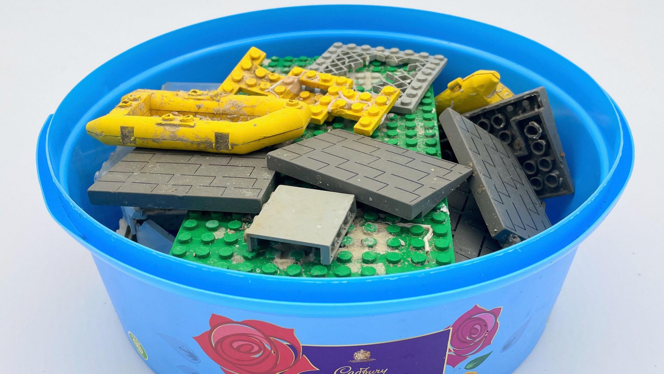 A blue plastic sweet tin full of found Lego pieces including two yellow life rafts, a large green model base and grey roofing tiles.