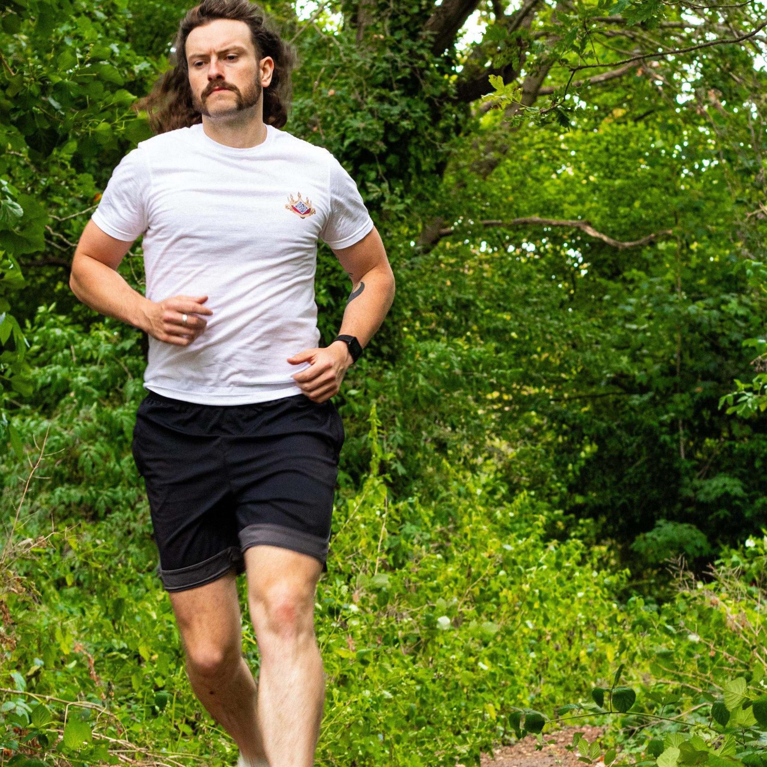 Iain Ward is seen with long hair and a moustache running along a leafy path. He is wearing a white T-shirt, black shorts, and a smartwatch, with green trees and plants surrounding the trail.