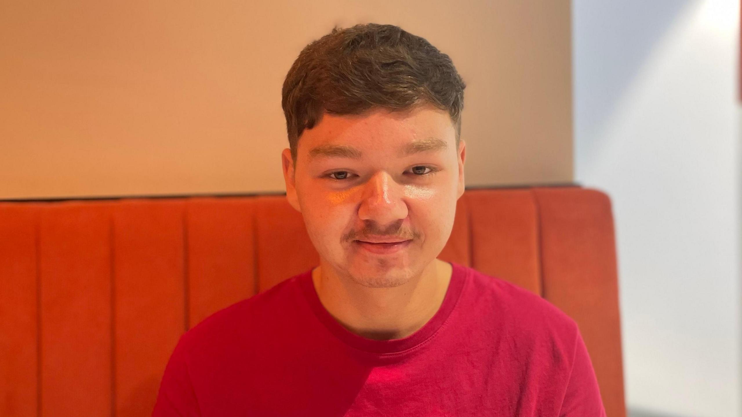 A young, male student with brown hair wearing a red t-shirt stares at the camera