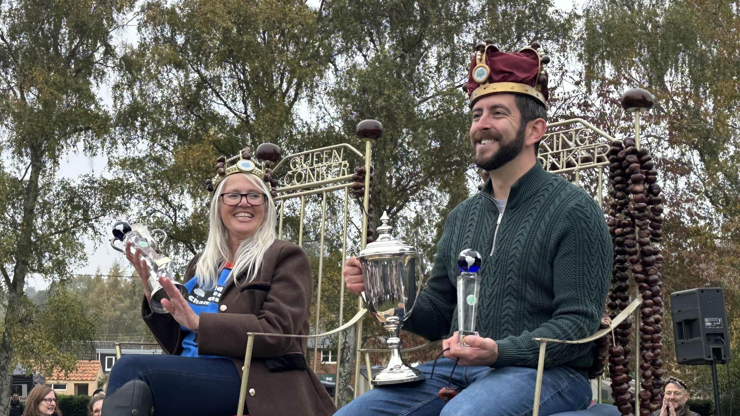 A woman and a man sitting on chairs that say Queen Conker and King Conker across the back. They are both holding silver trophies, wearing crowns and smiling. She has long grey hair and is wearing glasses, a brown coat and blue jeans. He has a dark beard and is wearing a green jumper and blue jeans.