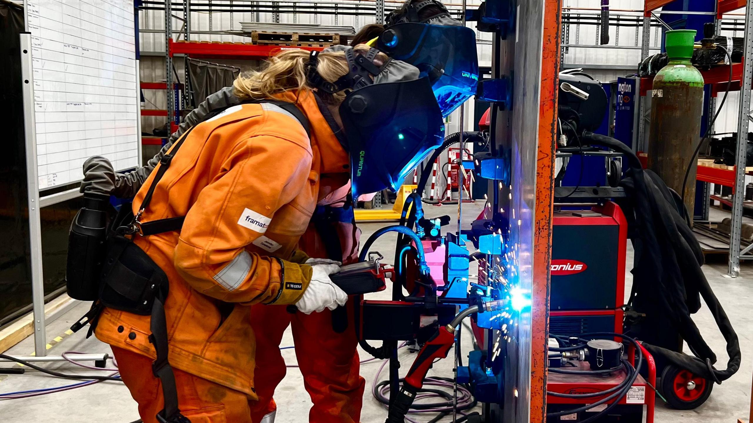 Two welders in full PPE with darkened visors stand in front of a sheet of steel. Blue sparks fly from where the welder is striking the metal.