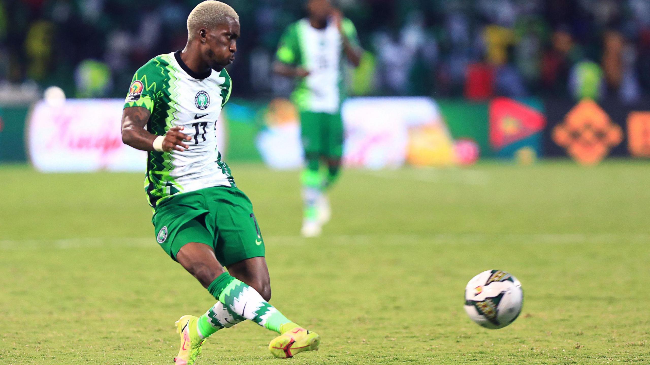 Henry Onyekuru with short bleached-blonde hair wears green and white Nigeria kit as he executes a side-footed pass of a football, which is seen in shot moving just above ground level, during a match at the 2021 Africa Cup of Nations