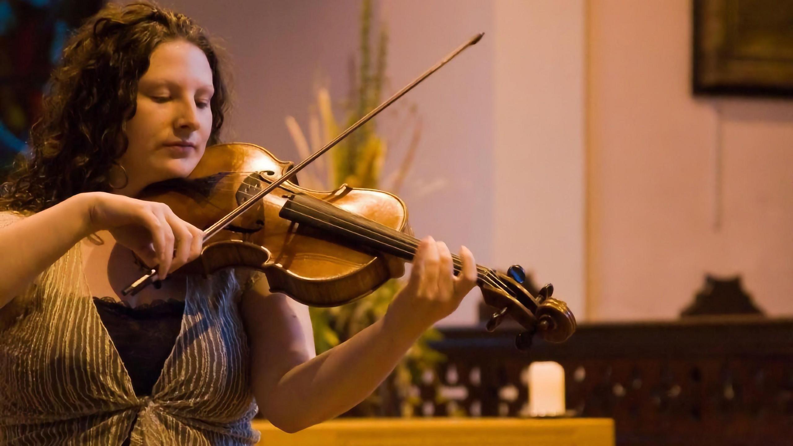 Laura Rowles, a woman with dark curly hair, looking down at the strings as she plays the violin.