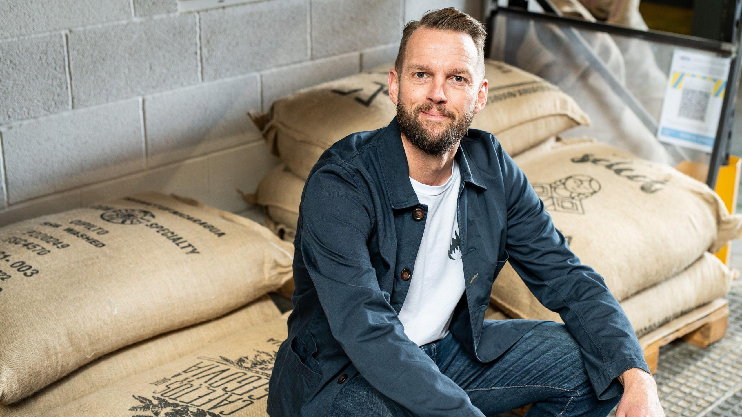 Will Kenney, smiling in open blue shirt, white t-shirt and jeans. He is sitting on bags of coffee beans in the company's roastery in Nottingham.