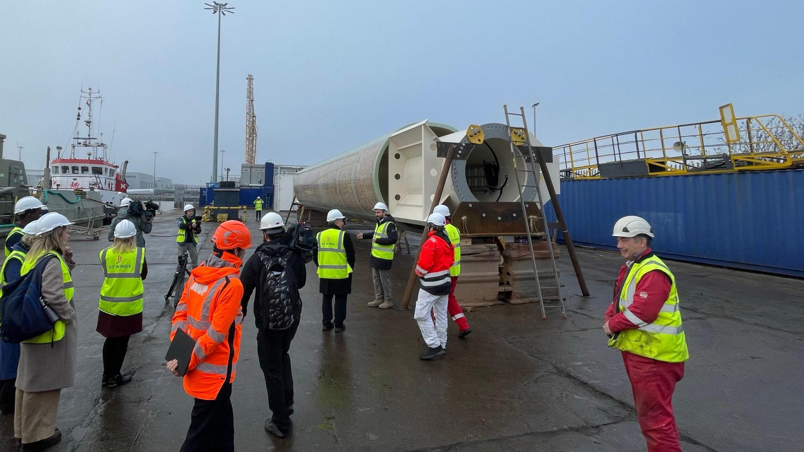 A group of people wearing hard hats and hi-vis vests standing on a dockside looking at a large tube-like structure. A ship is in the background