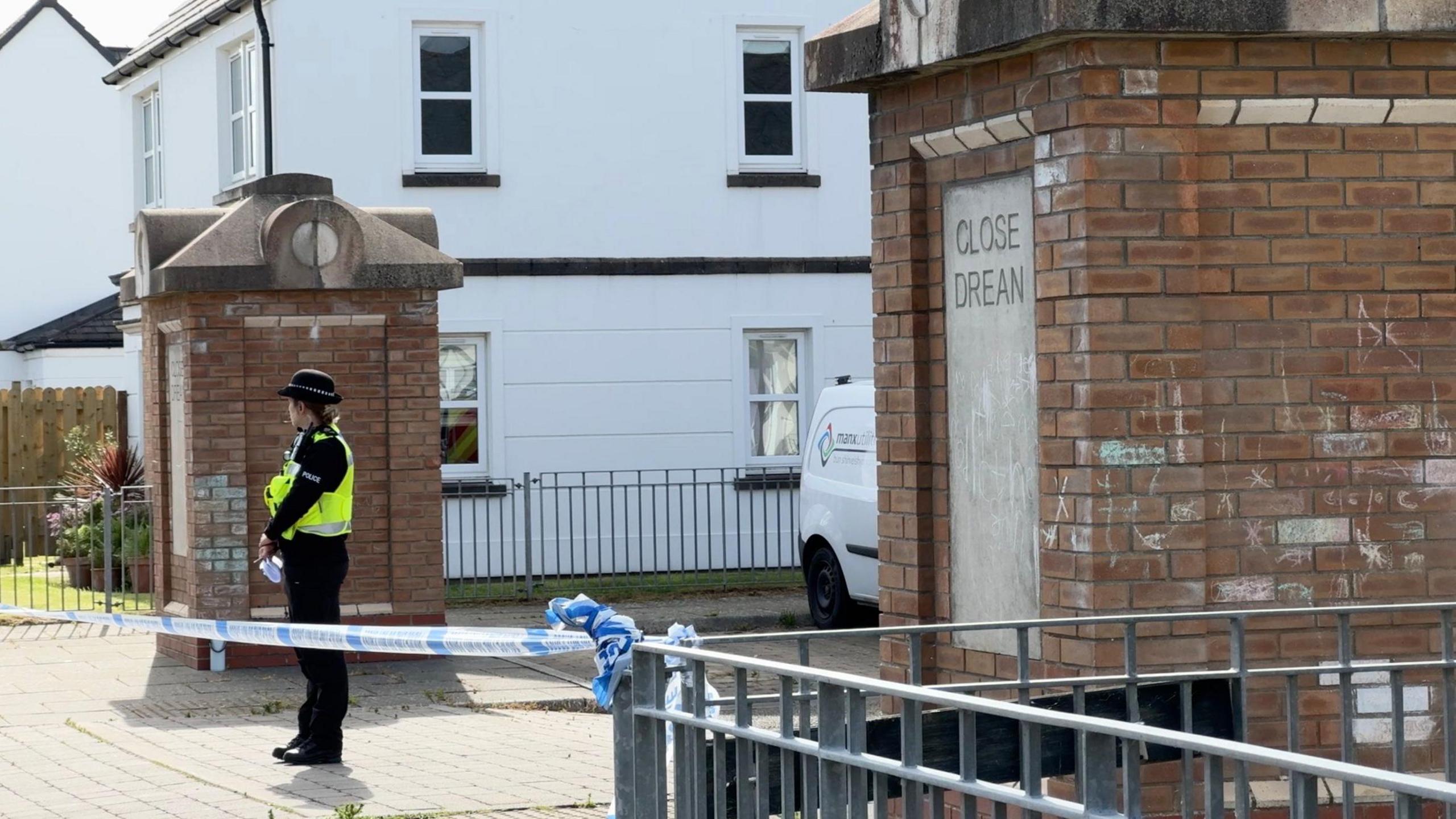 A policewoman stands at the entrance of Close Drean behind police tape cordoning it off.