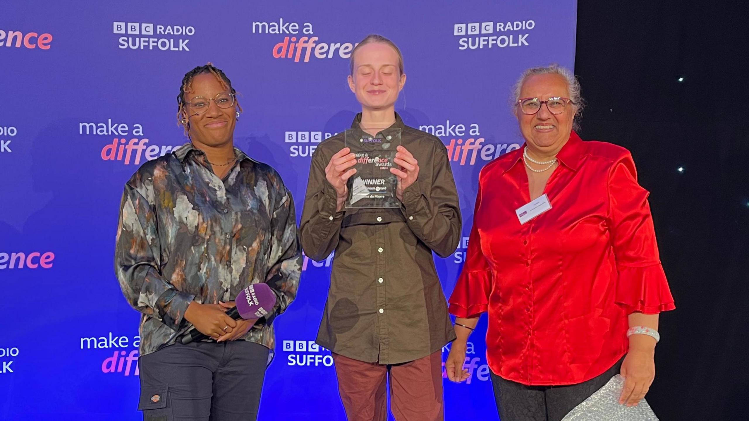 Angelle Joseph, James de Mierre and Gulshan Kayembe stand on a stage. James is holding a glass trophy. The backdrop says BBC Radio Suffolk Make a Difference