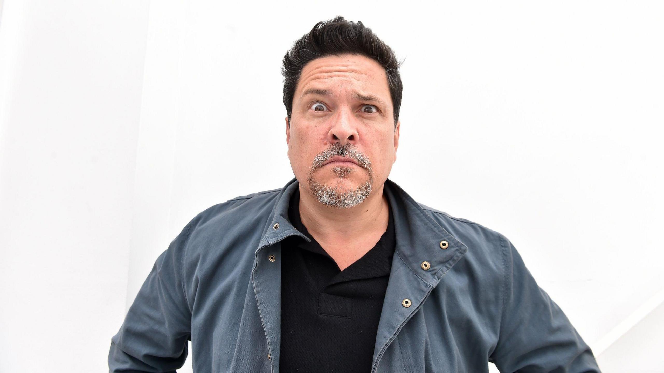 Dom pulls a confused face at the camera in front of a white background. He has a grey blue shirt on with a black top underneath. He has brown eyes with short black hair. He has a grey goatee as well.