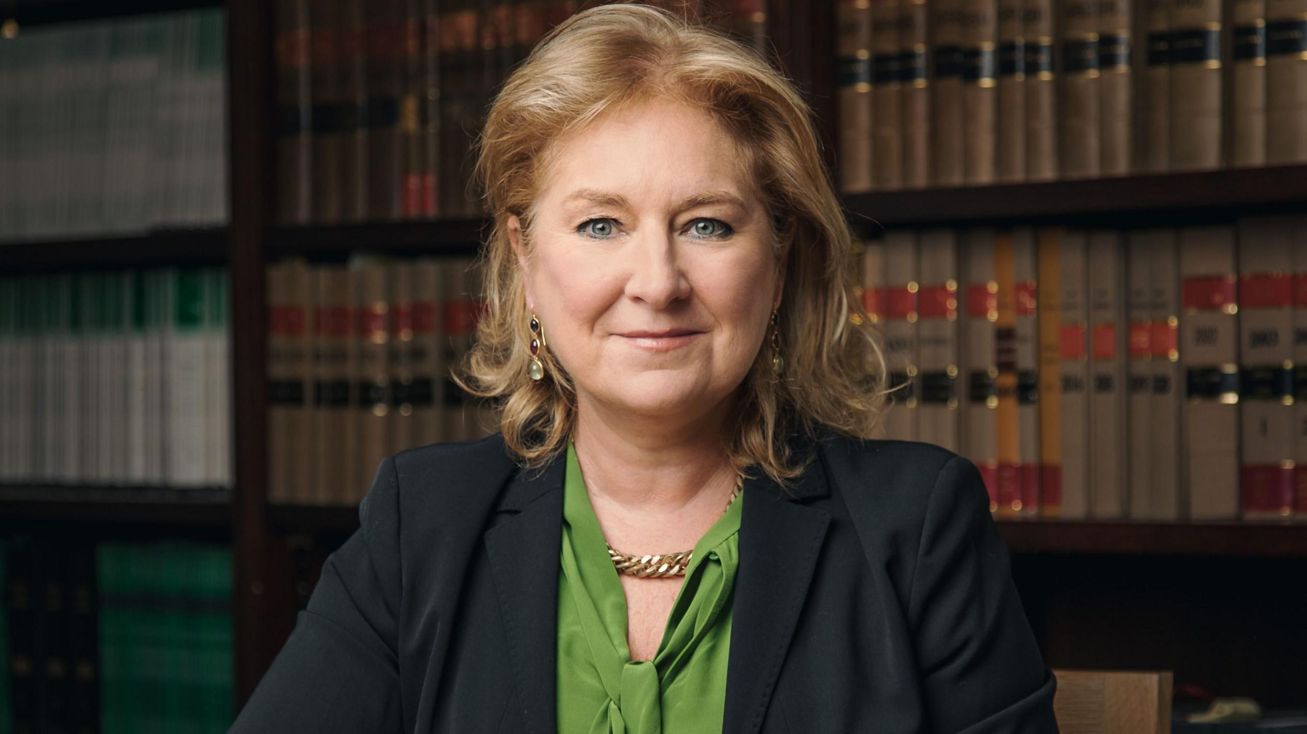 Baroness Sue Carr sits in front of a bookshelf with her arms crossed. She's wearing a green shirt and black suit jacket. 