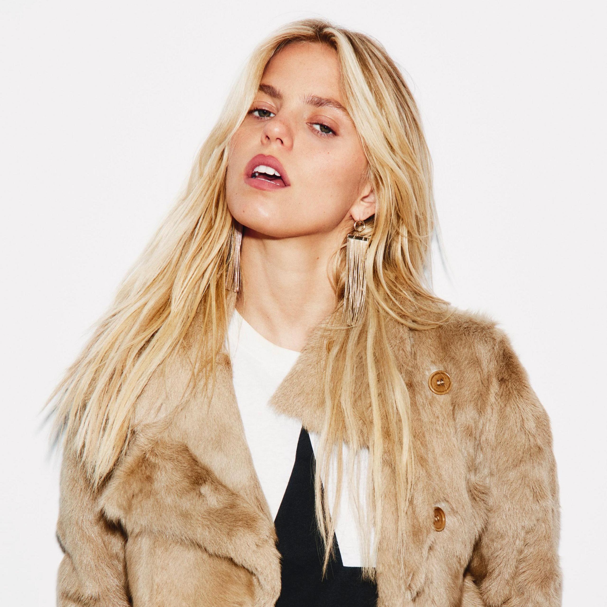 Reneé Rapp poses against a plain white backdrop. She is wearing a white t-shirt with a bold, black star in the middle, covered by a shaggy faux-fur jacket.