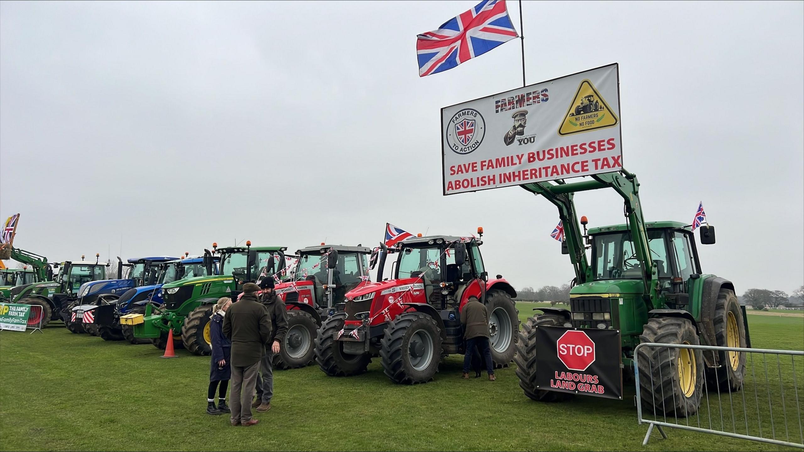 A green tractor - at the start of a line of them - holds up a white banner saying: Save Businesses, Abolish Inheritance Tax. A group of three people are standing in front of the tractors, parked on grass.