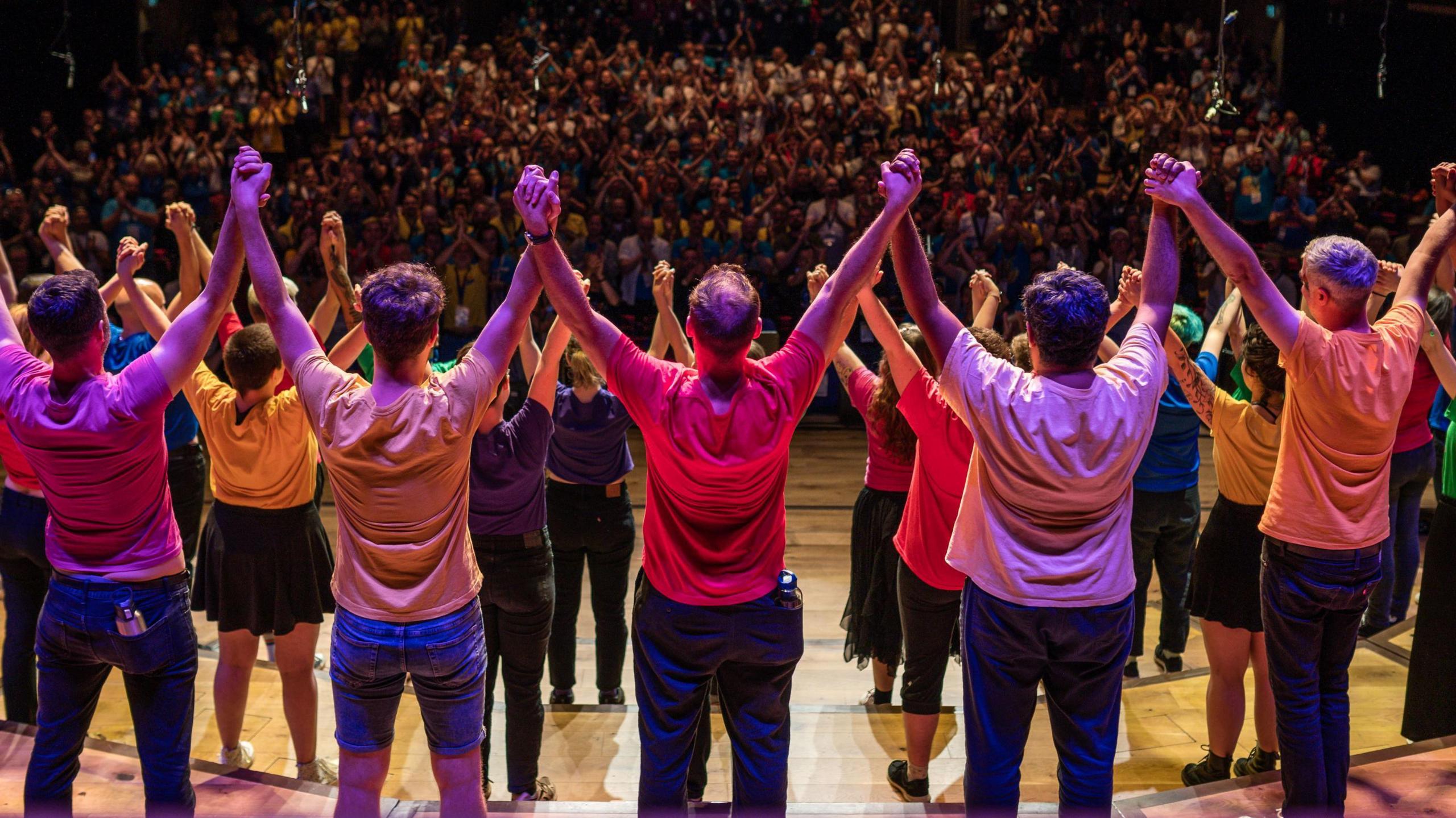 Dozens of people in colourful T-shirts stand on a stage at the Bristol Beacon, linking hands as they salute the audience who are applauding in the background