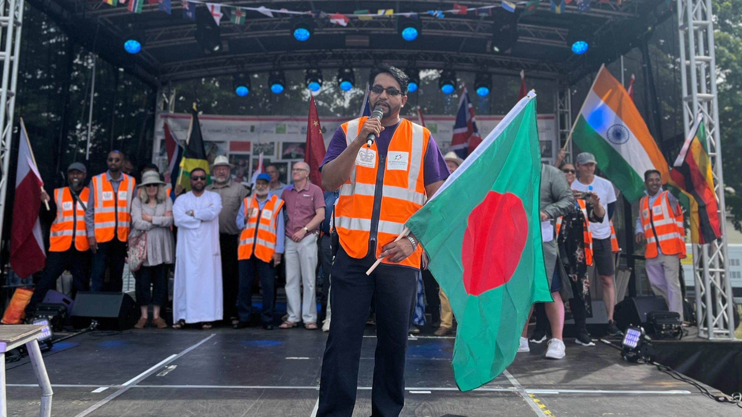 Boshor Ali, a man of Bangladeshi heritage, holds a green flag with a red circle in the centre as he stands on a festival stage.