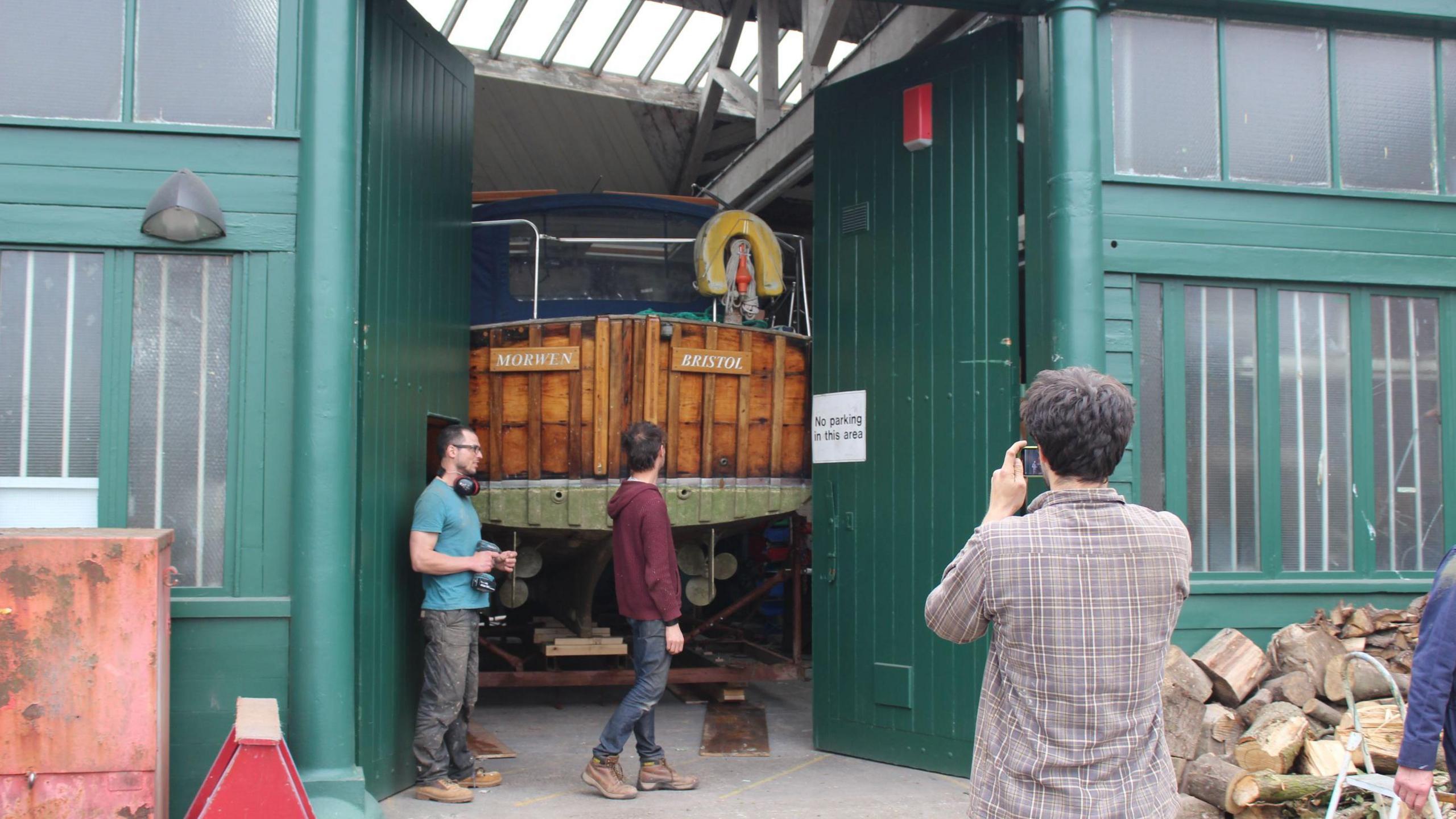 The entrance to the Shipwrights building. It is a large wooden industrial shed which has been painted a dark green. The doors are open and there is a wooden boat backing out, with two men stood beside it and another taking a picture as it comes out. 