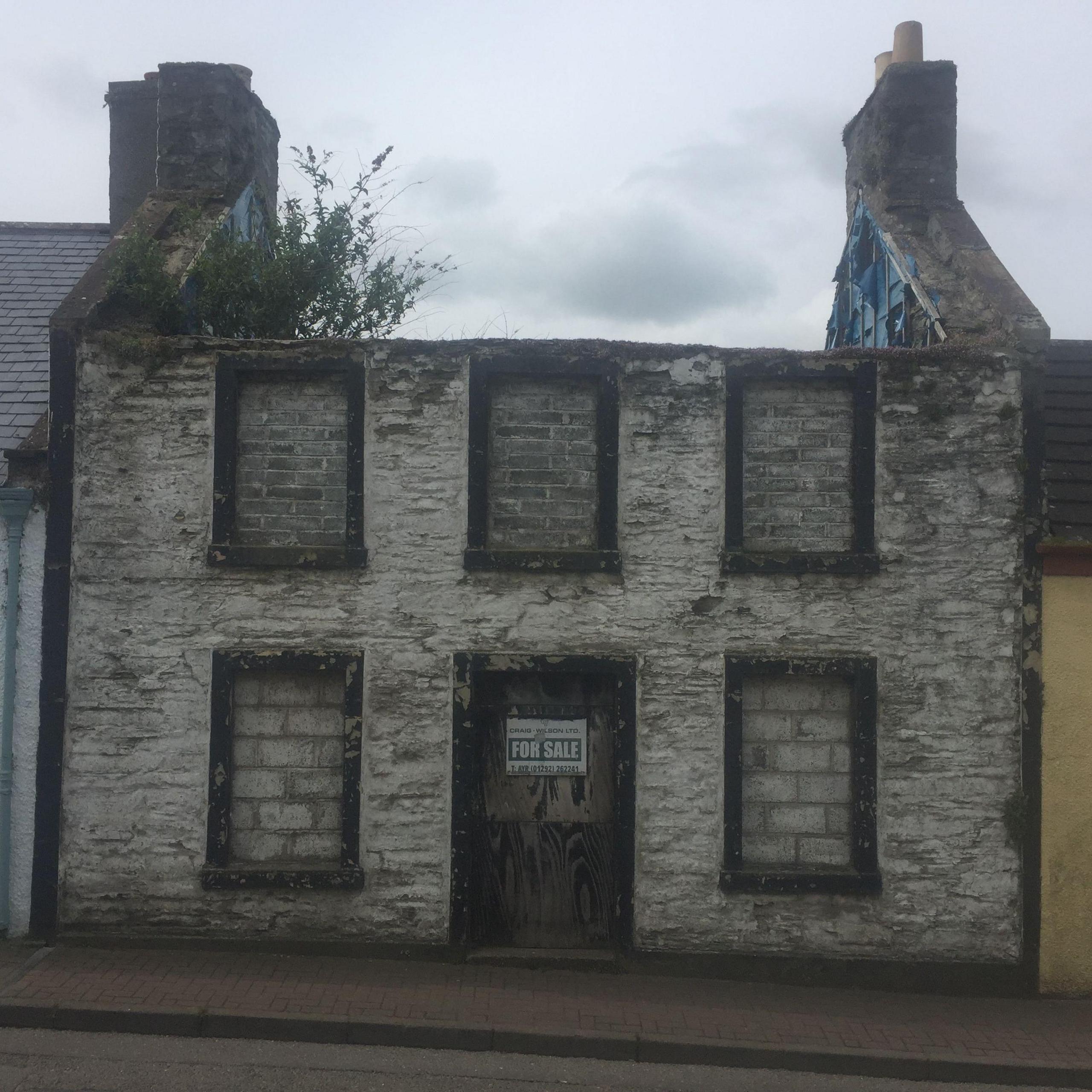 A decaying stone building with dirty and molding bricks and filled in windows, there's no roof and a tree is growing out the top of the property.