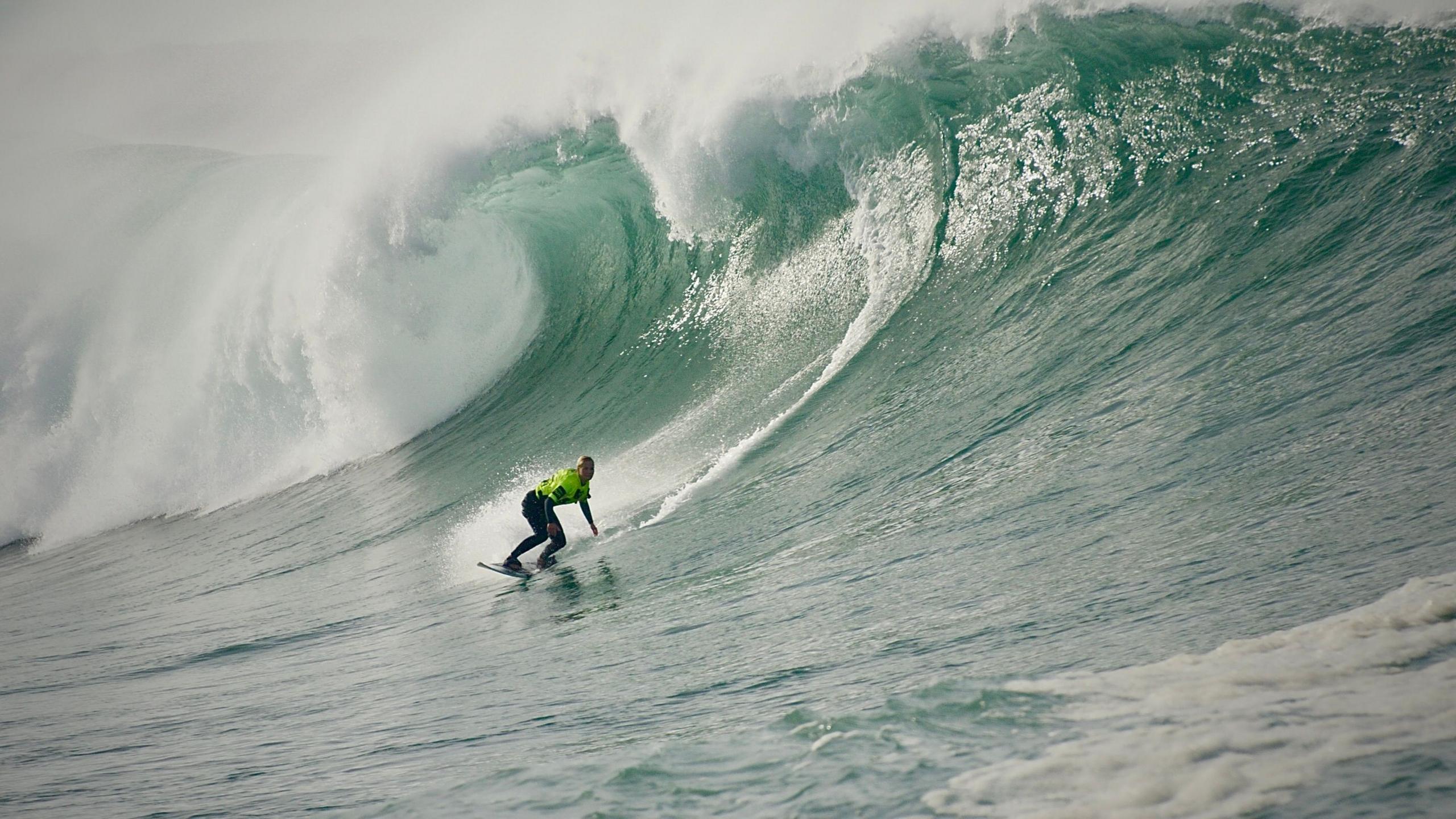 Robyn surfing a huge wave. The wave is curling over. Robyn is wearing a bright coloured wetsuit.