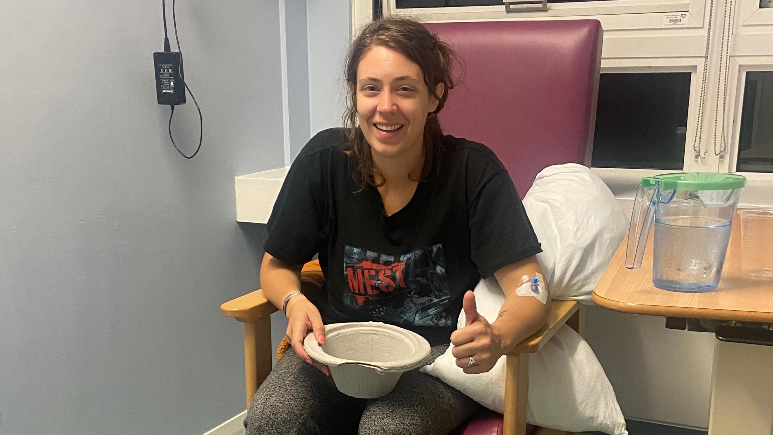 Kat is wearing a black t-shirt and is sitting in a chair. She is holding a cardboard sick bowl and has brown hair.
