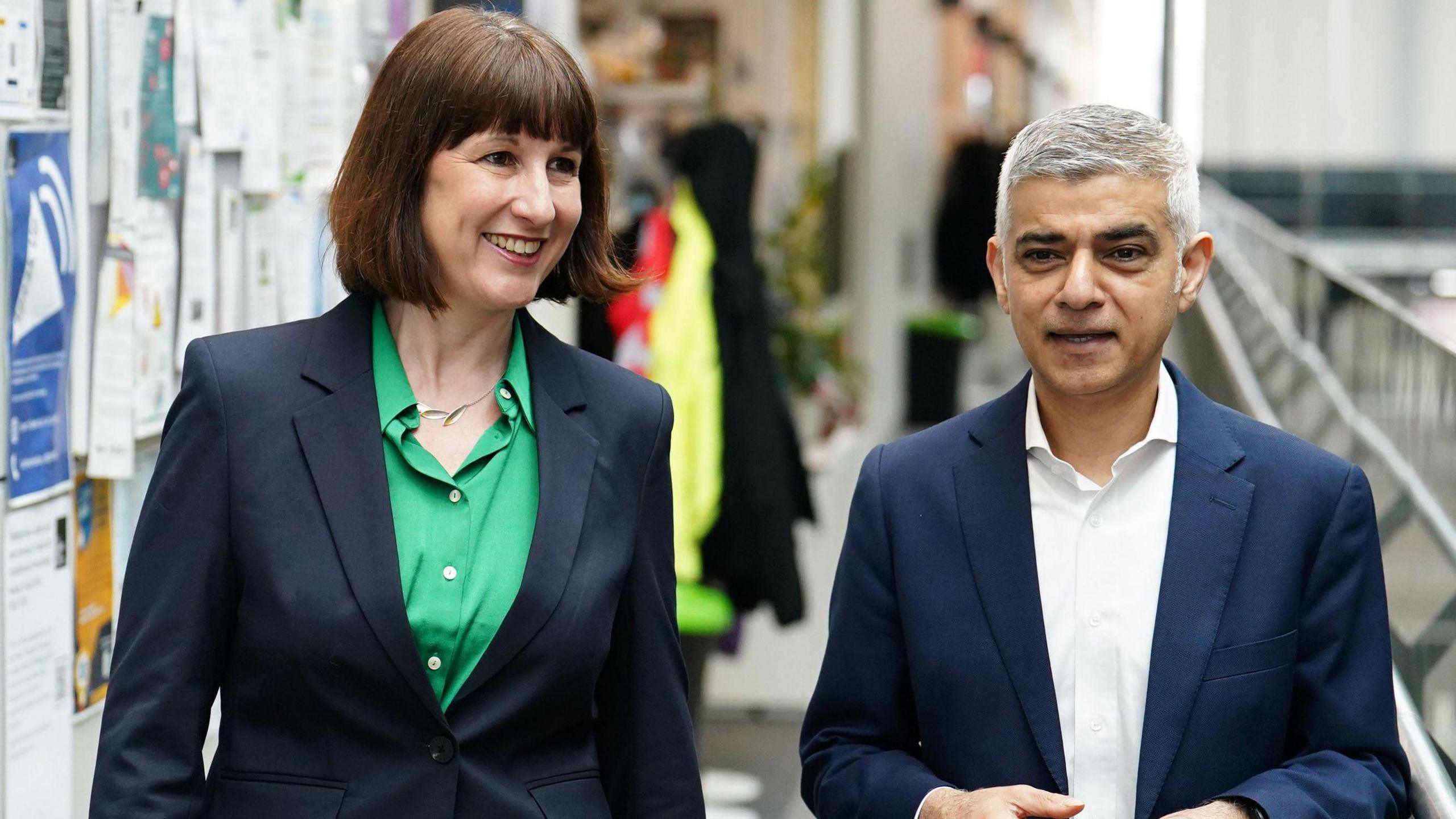 A smiling middle aged woman in a dark jacket and green blouse standing next to a man in a dark blue suit jacket and white shirt. They are walking down a corridor. 