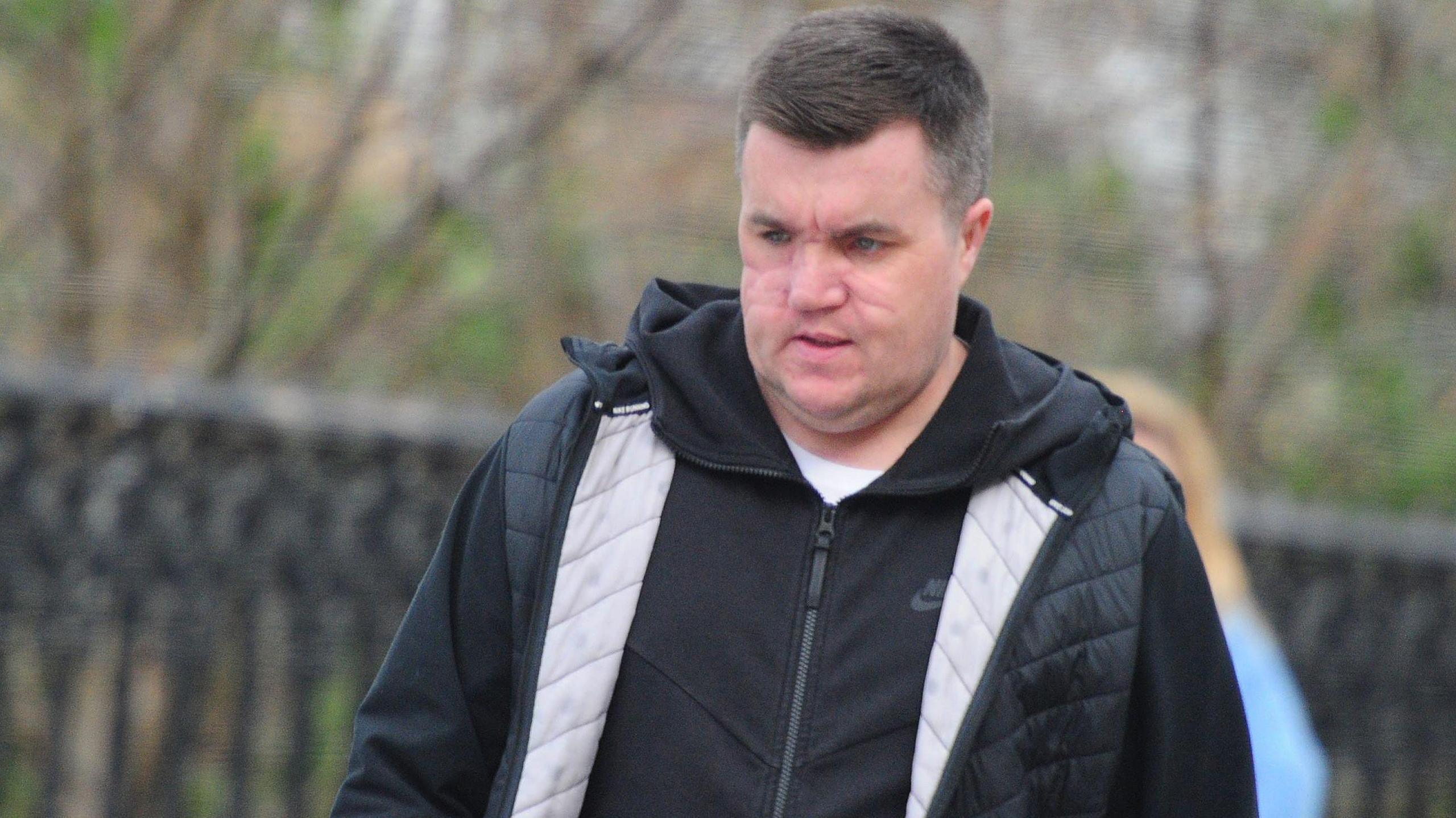 Steven "Bonzo" Daniel pictured walking along a street, with a black fence and trees visible but blurred in the background. He is wearing a black hooded jacket and Nike jacket and a white t-shirt. He has short black hair, combed to the side and is looking at the ground. He has extensive facial scarring.