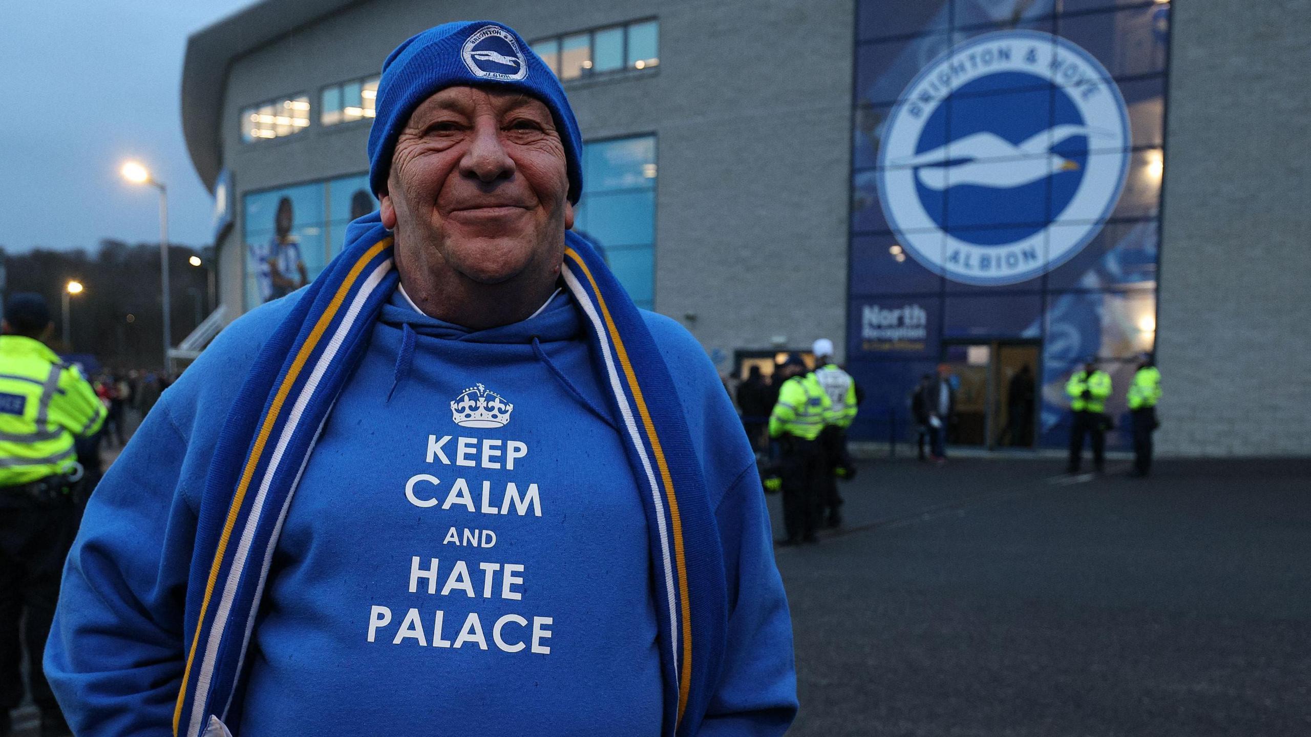 A man in a Brighton and Hove Albion beanie and hoodie that says "Keep calm and hate Palace". He is standing in front of a Brighton badge on the outside of the Amex.
