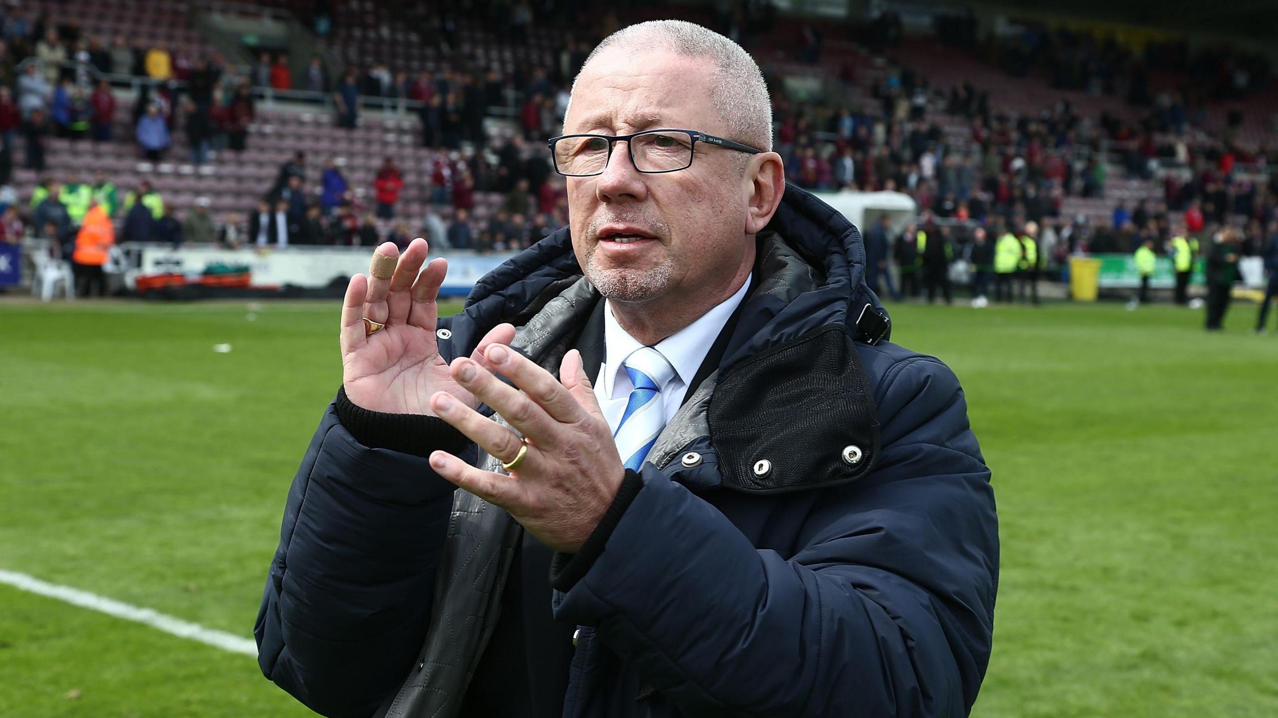 Paul Scally, wearing a white shirt and Gillingham FC blue and white tie and a blue anorak, applauding fans at the Priestfield football ground in Gillngham.