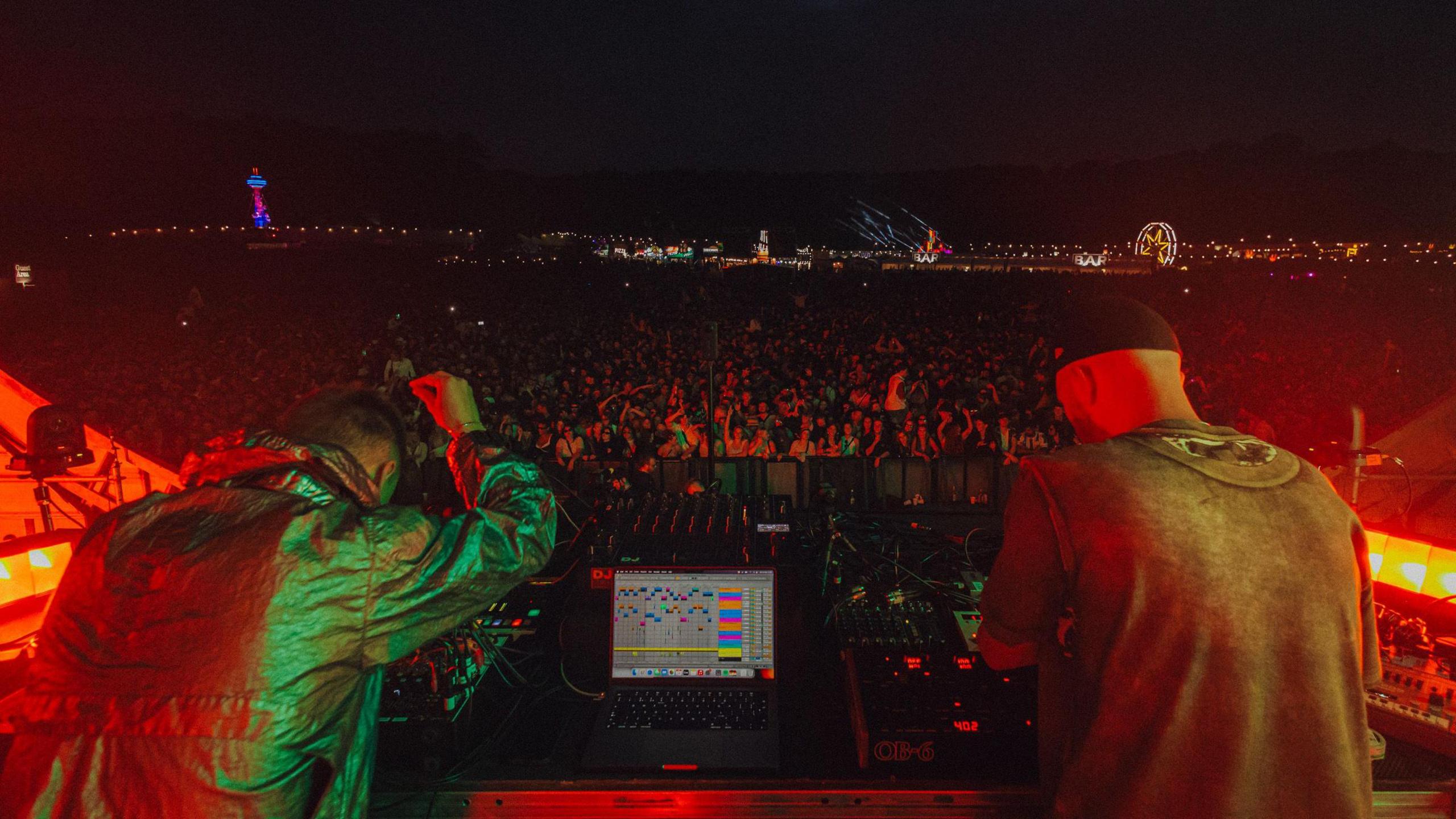 A view looking out behind two DJs, with a laptop in front of them, to the crowd beyond at night.