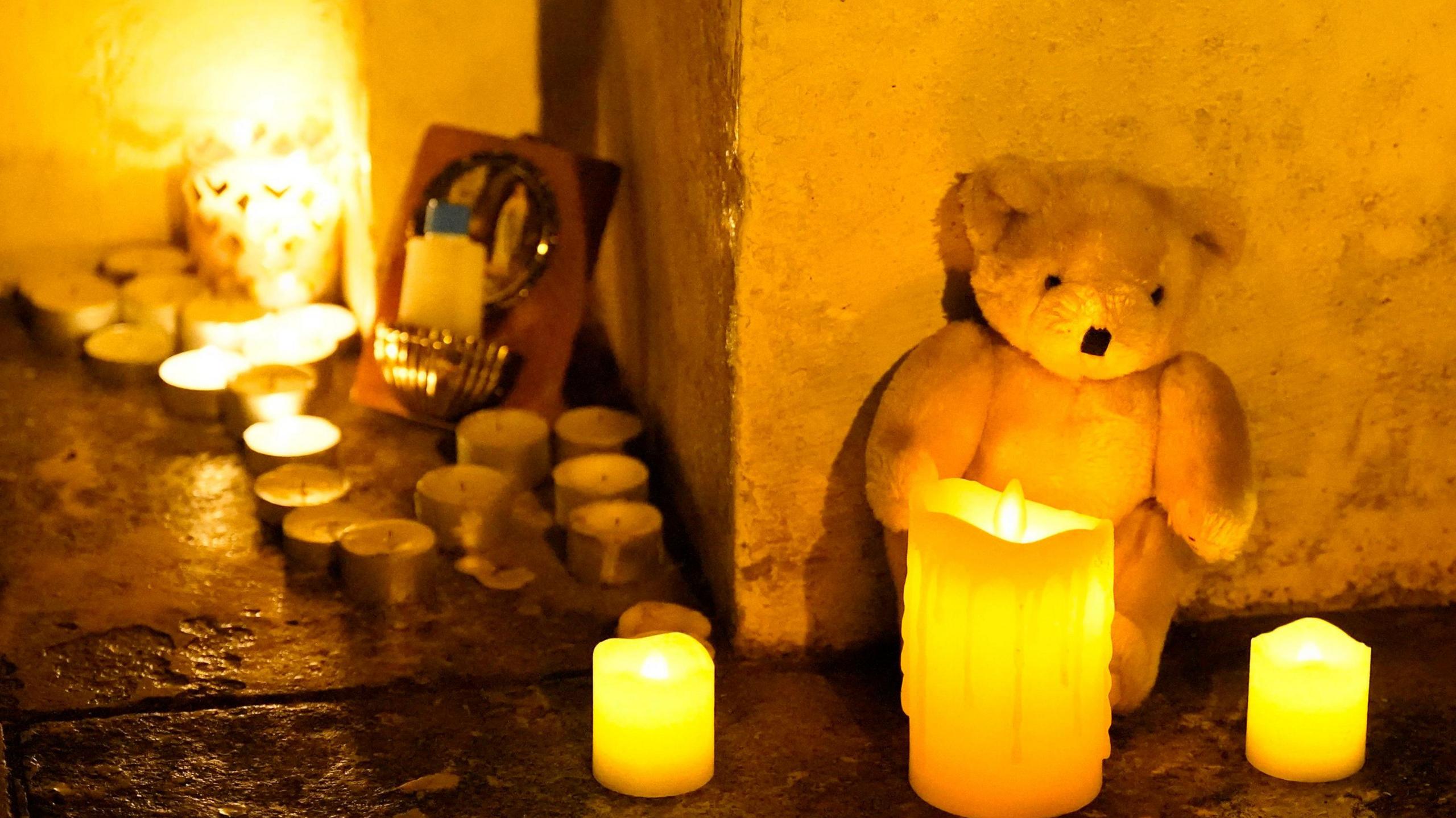 A white teddy bear sits against a cream wall with three candles in front of it. There are a collection of about a dozen tealights to the right of the teddy.