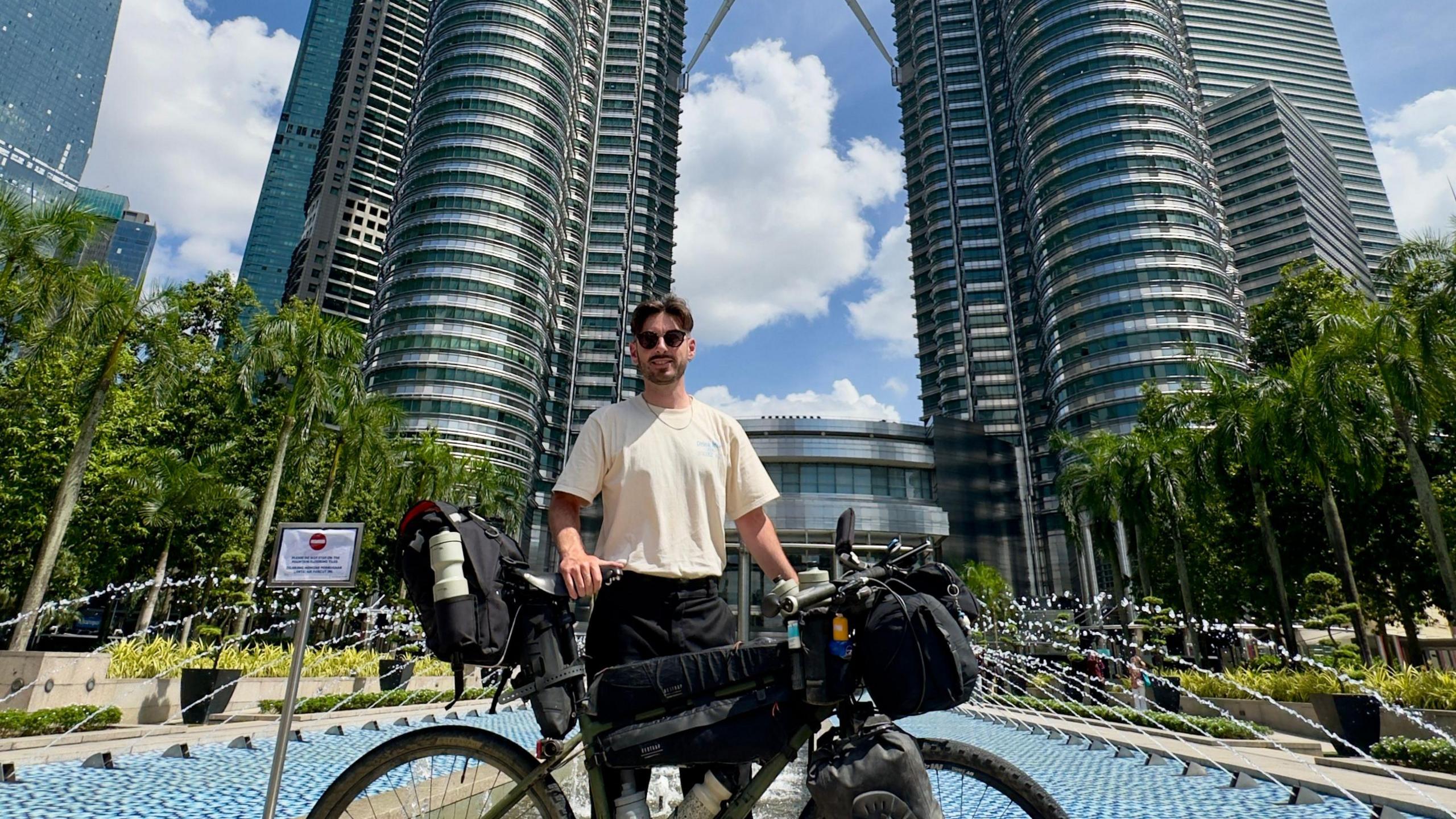Man with dark hair and sunglasses poses with his bike in front of the Petronas Twin Towers in Kuala Lumpur, Malaysia
