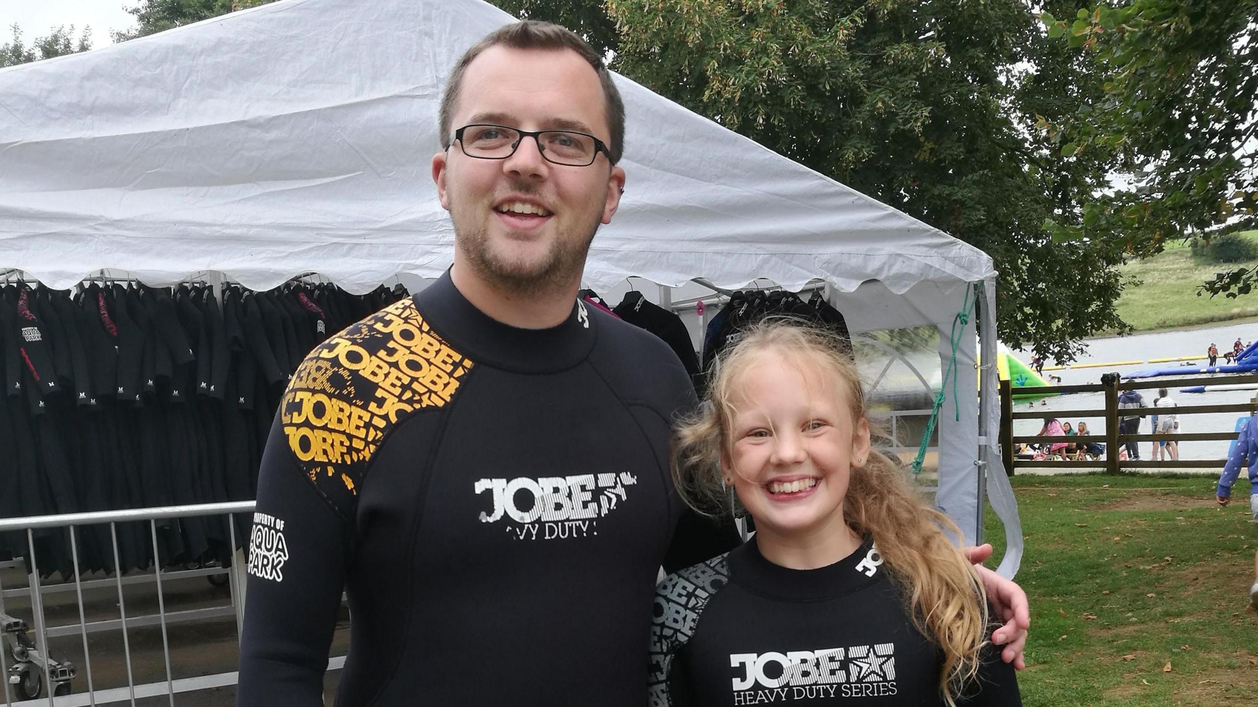 A man with short brown hair, stubble and rectangular glasses smiling wearing a black wetsuit. He has his hand around a young girl with curly blonde hair tied back who is giving a big smile and also wearing a black wetsuit. A marquee can be seen behind them as well as a lake and people dotted about to the right.