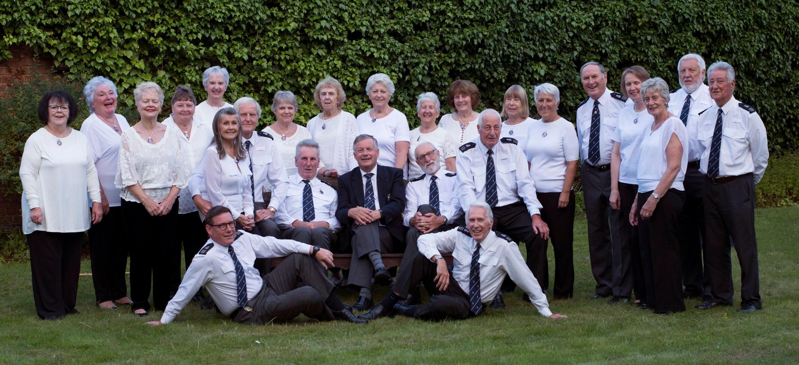 26 members of the choir, most wearing white tops.  Most are standing on a lawn with some sitting down.  Green foliage in the background.