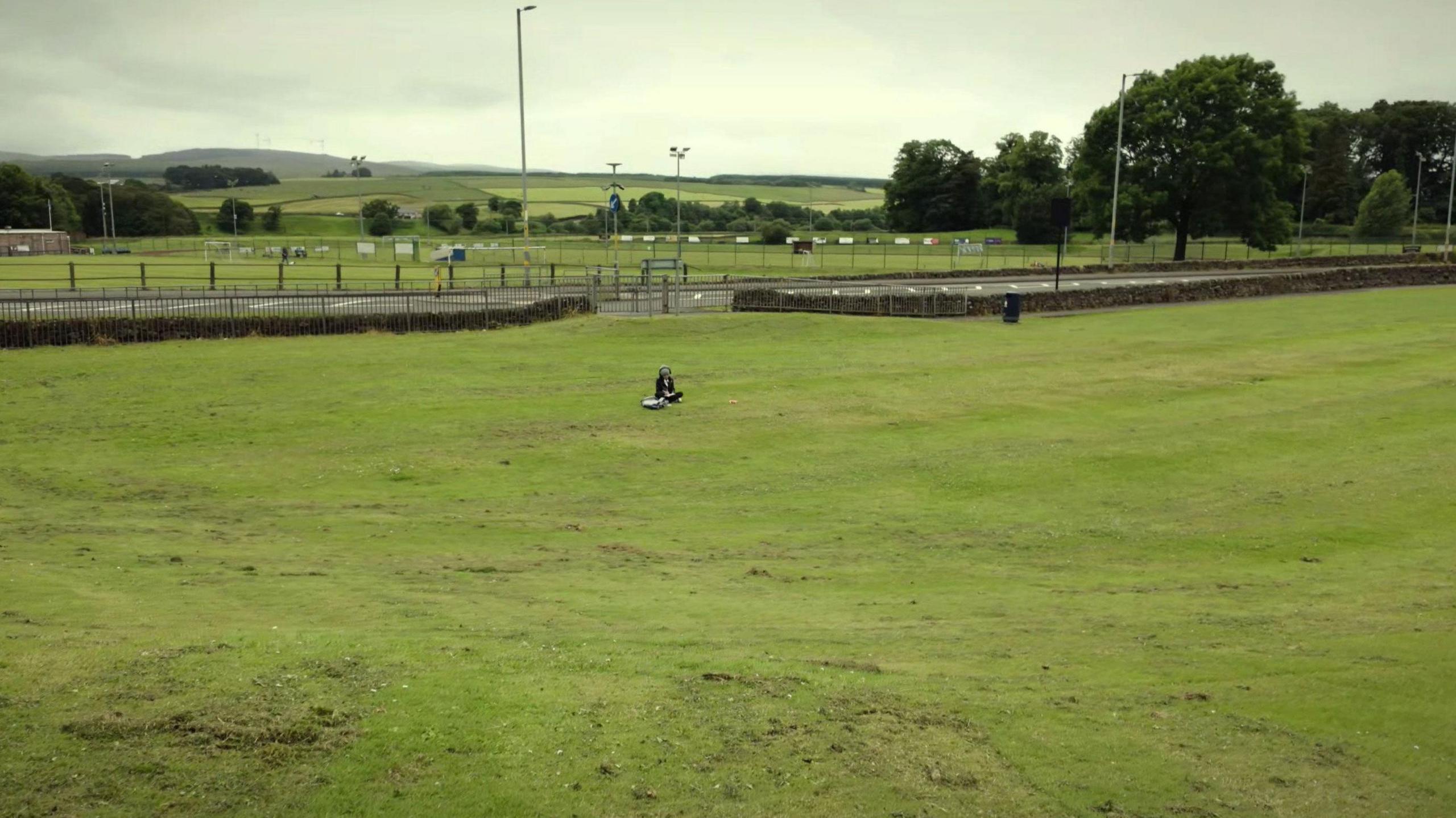 A scene of one young child in a large field sitting on their own surrounding by hills, trees and a nearby road