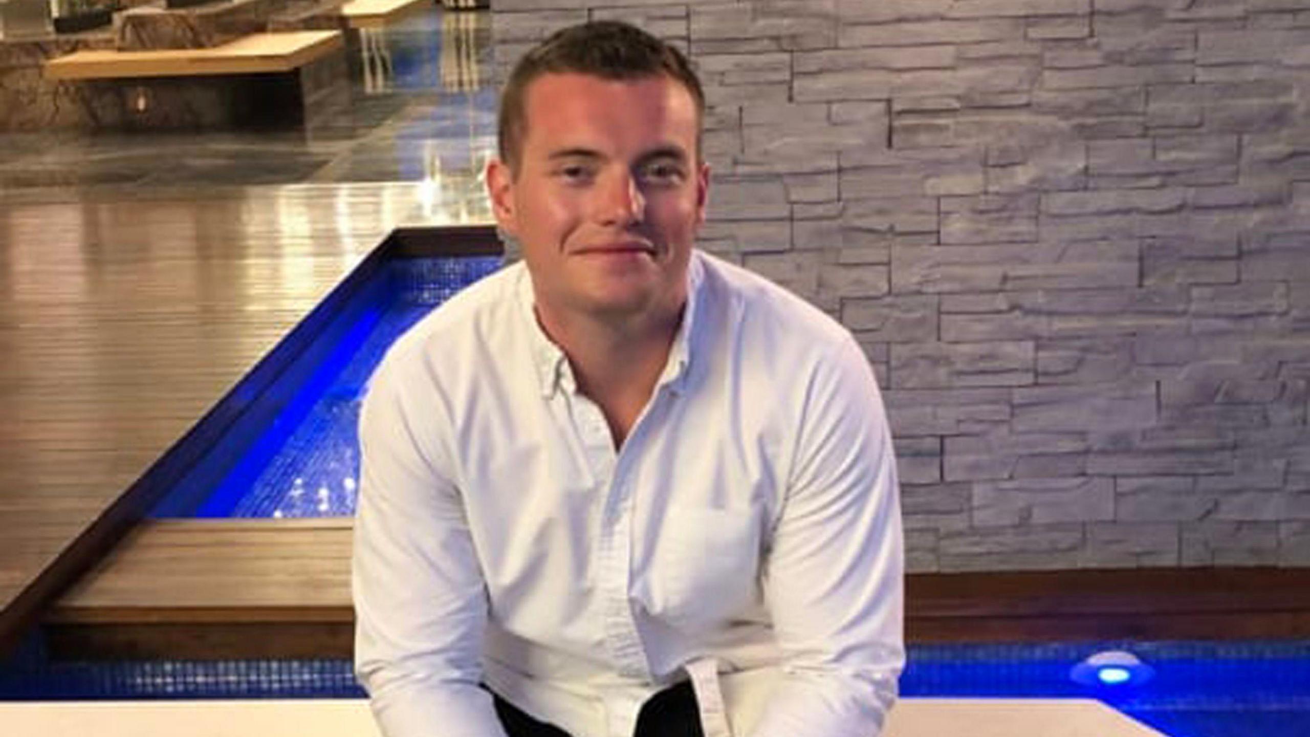 Jack Merritt pictured sitting at the edge of an indoor pool on holiday. It is evening and he wears a white shirt and looks happy and sunburned.