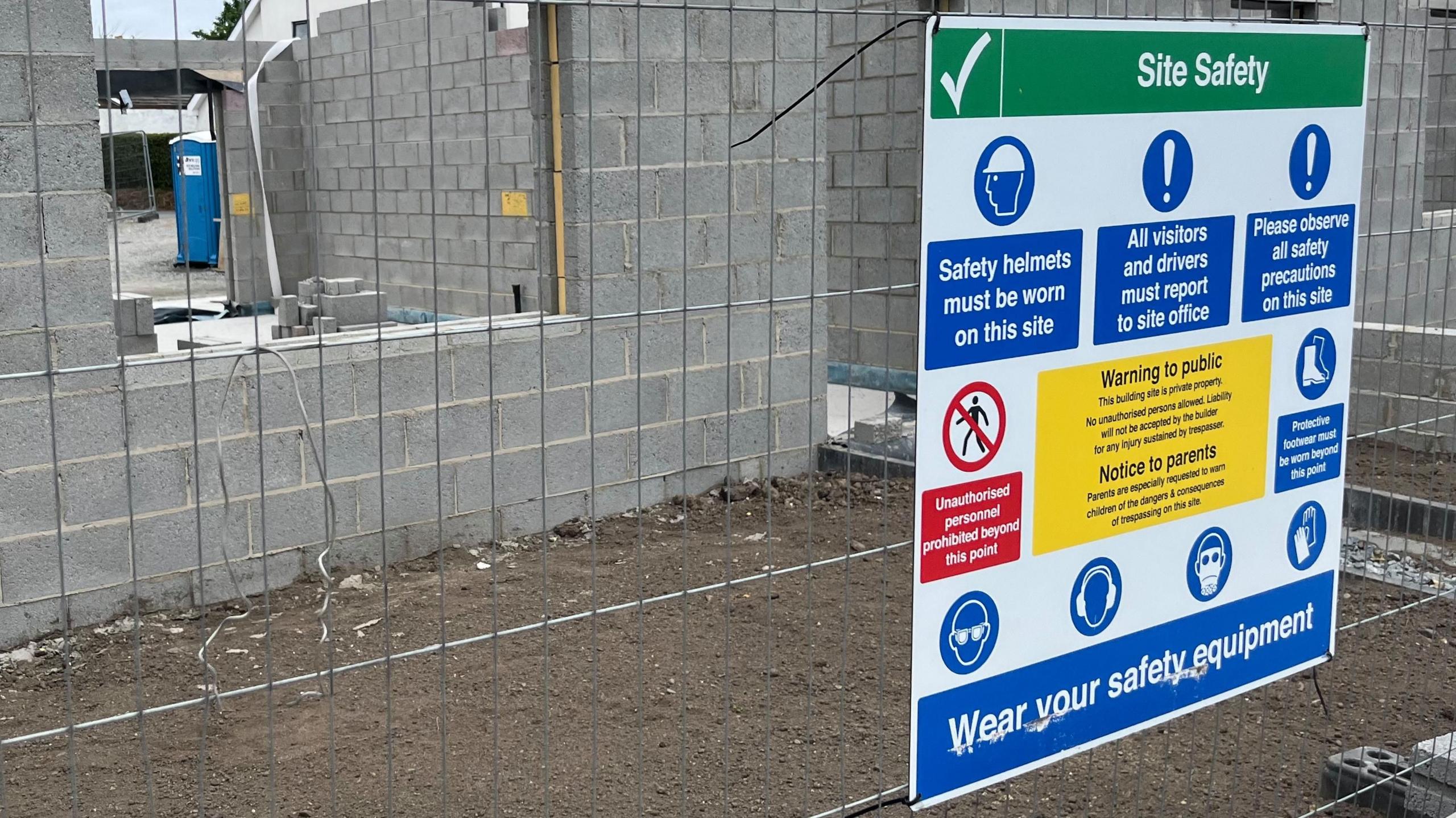 Image of a construction site safety sign with a a part-built, grey-brick building behind a fence. There is a sign on the fence giving guidance on site safety.