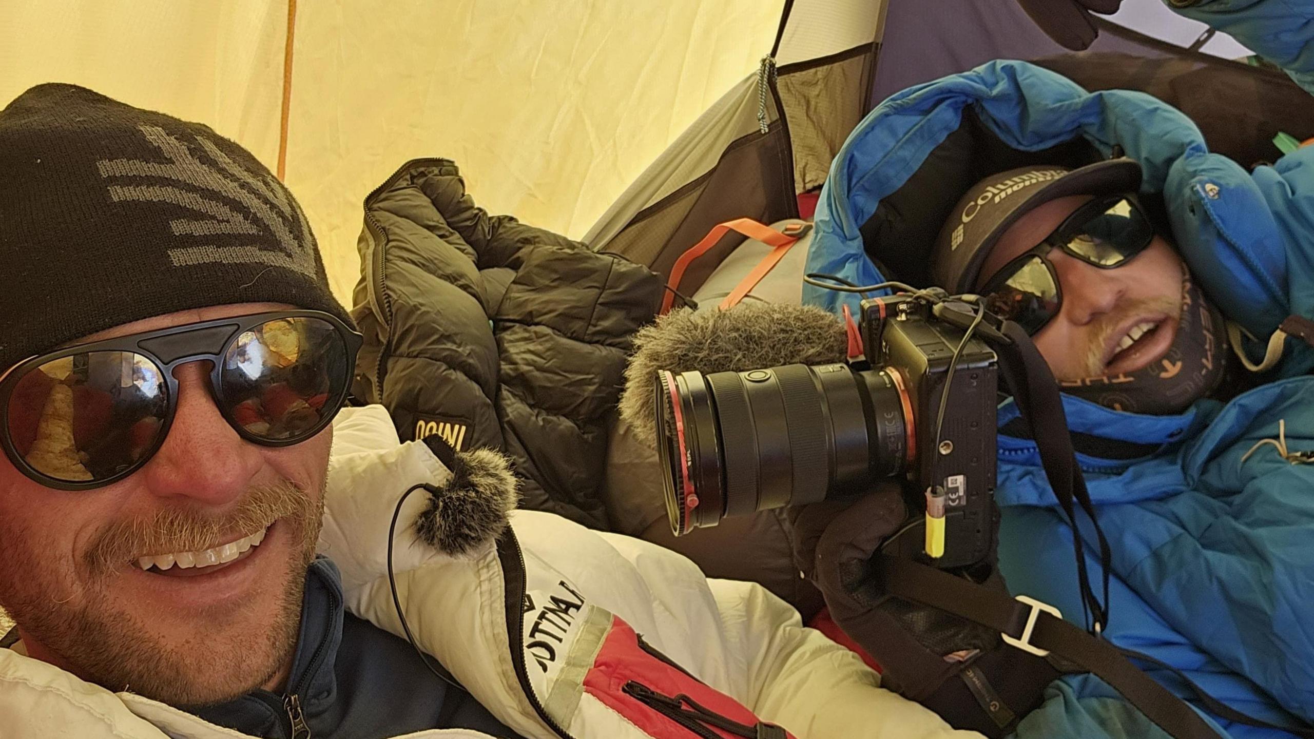 Two men in jackets and sunglasses are lying down in a tent. One of the men is holding a camera.