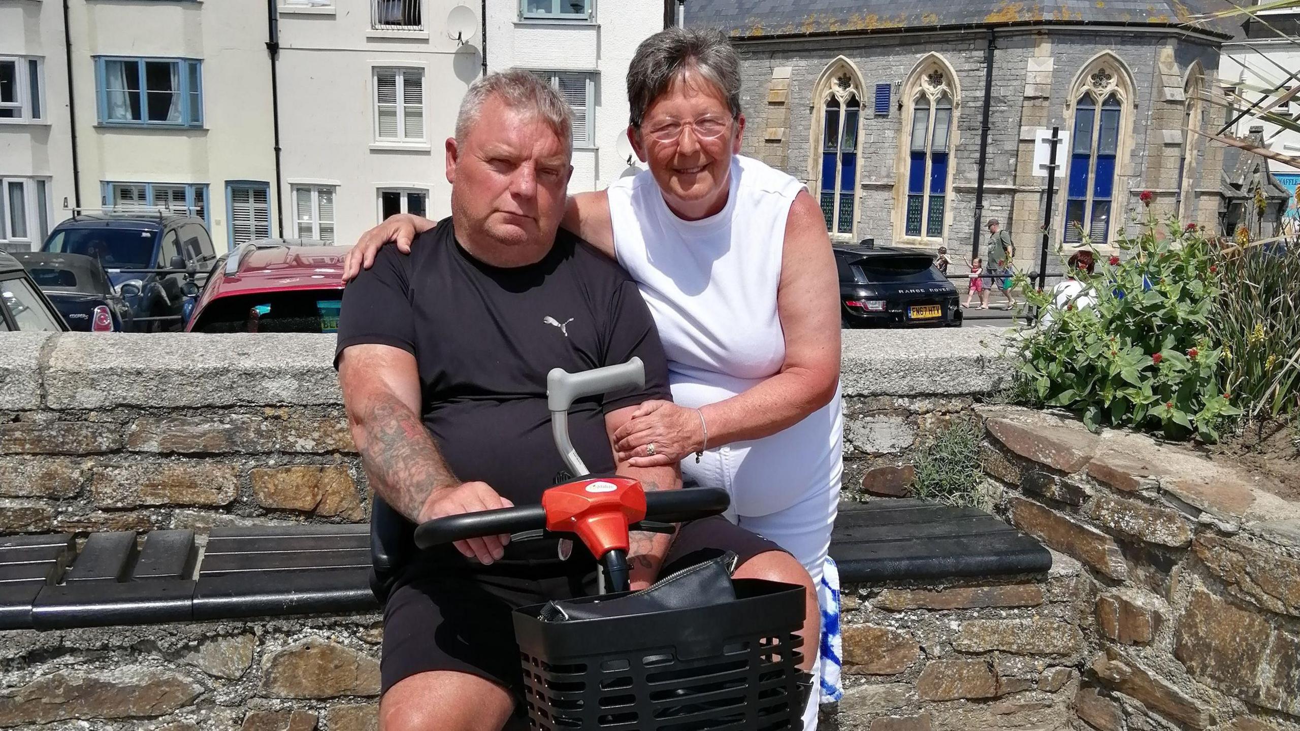 Wendy and Paul posing in front of a stone wall that has a road with cars, some houses and a church-style building behind them. Paul is wearing a black T-shirt and is sat on a black mobility scooter and holding his walking stick. Wendy is wearing a white T-shirt and trousers and is stood next to to him with her left arm on his shoulder.