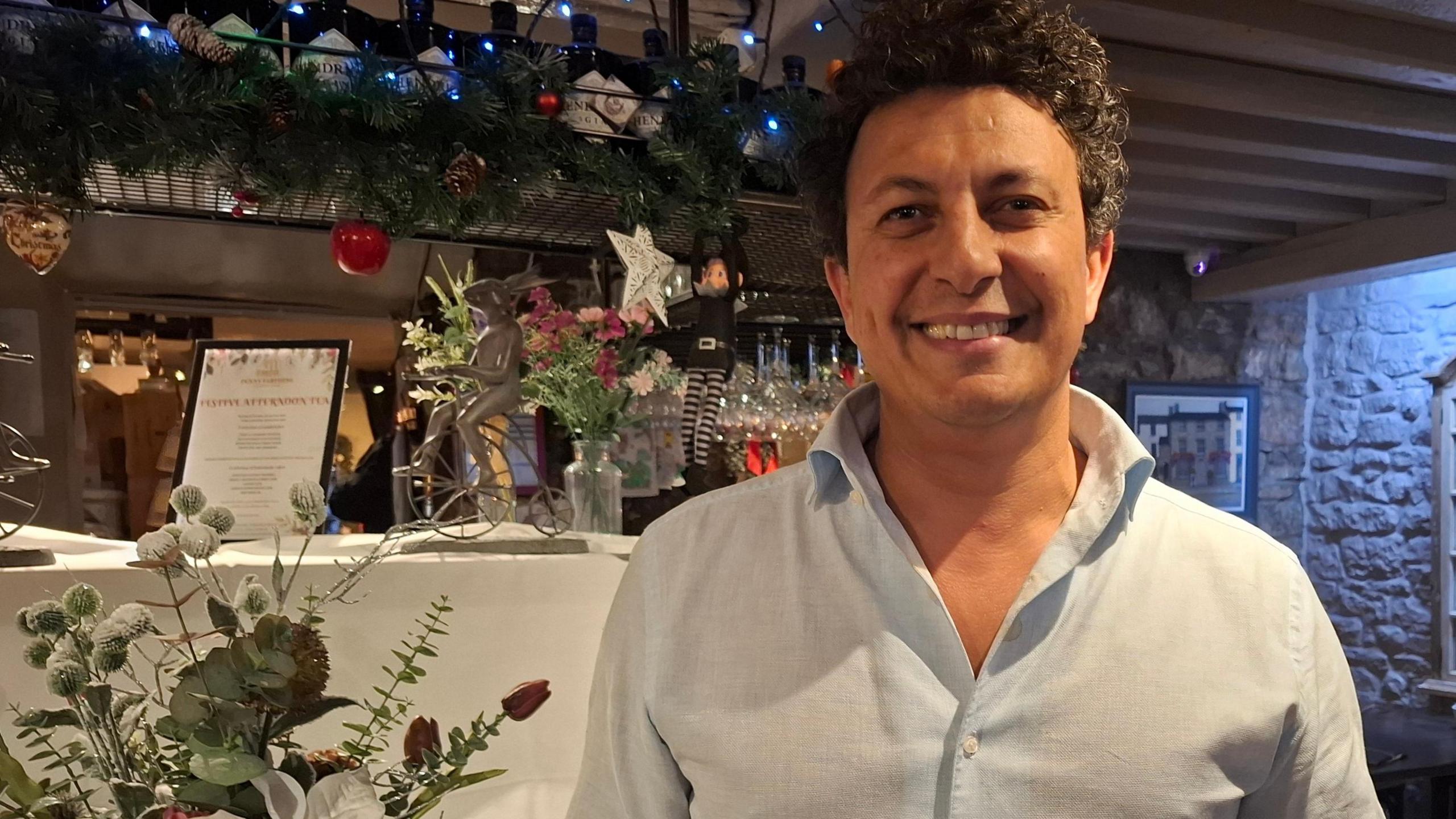 Mohammed Elkilay in the restaurant smiling and wearing a light blue shirt. he has dark curly hair and is standing in front of the bar