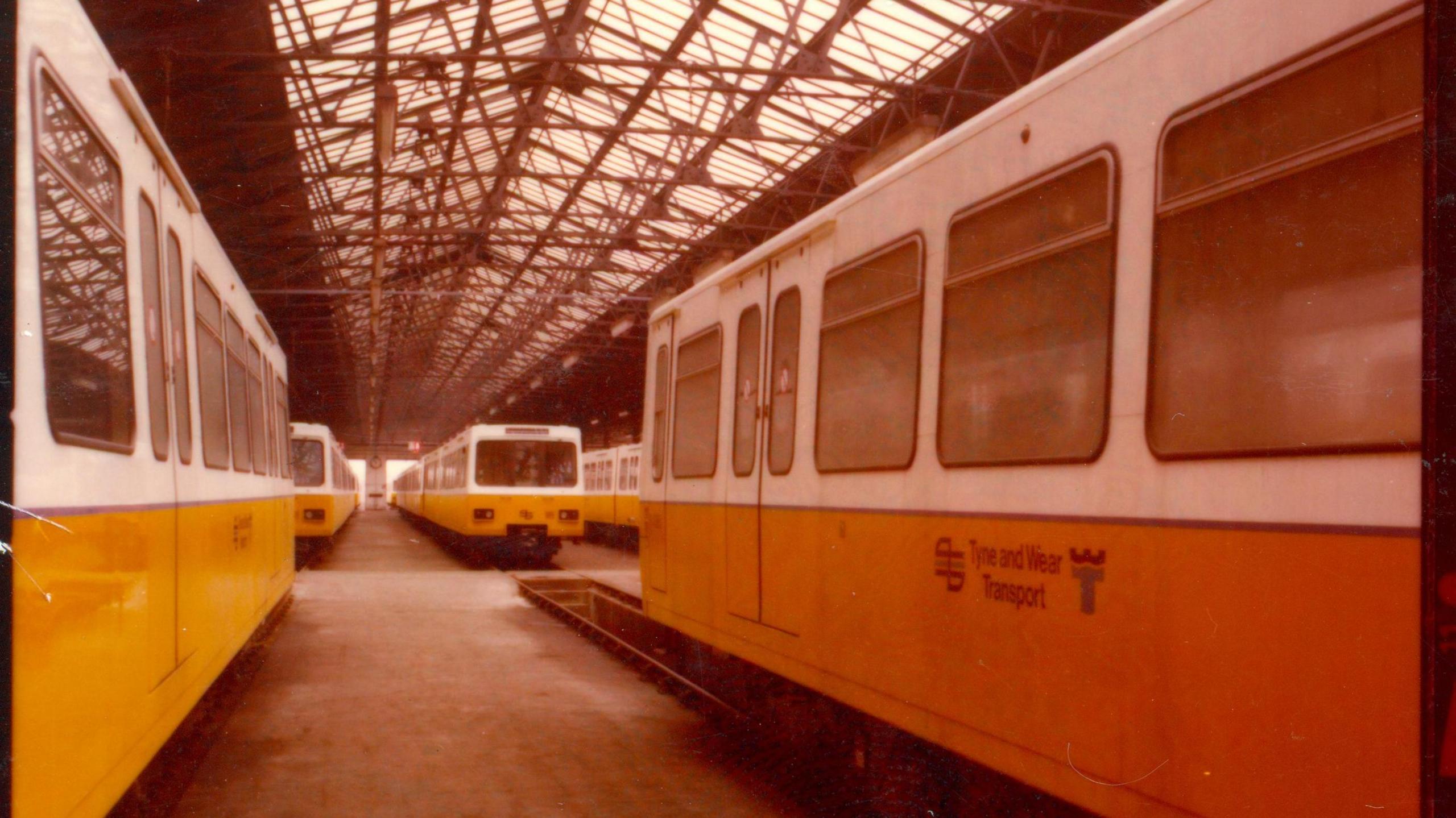 There are five original Metro trains, with yellow and white colouring, that are stationary in what appears to be a warehouse. There is a slight grainy quality to the image.