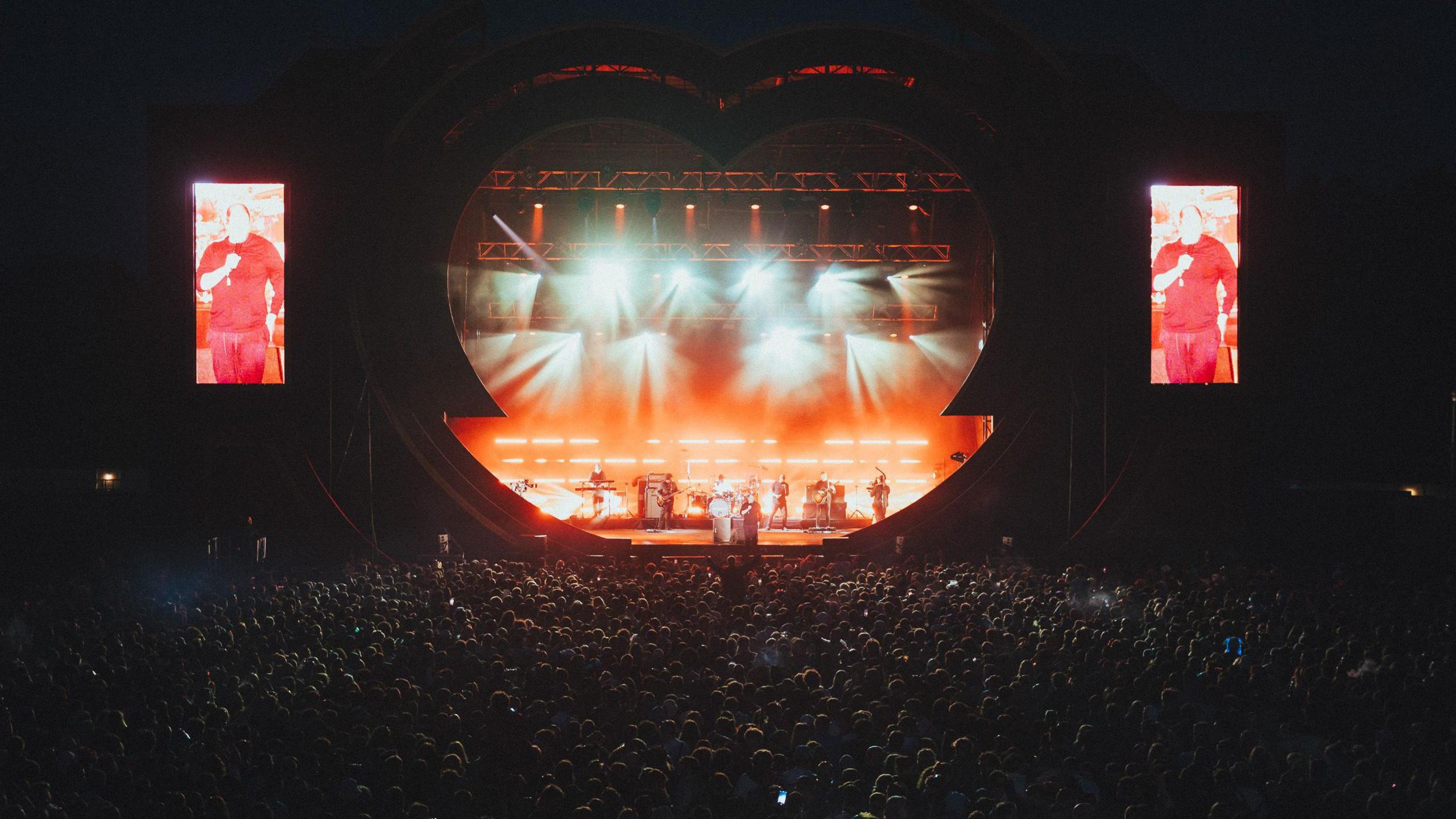 A large crowd is packed in front of a large heart-shaped main stage at the Love Saves The Day festival in Bristol in May 2024. The stage is illuminated with white lights as the band The Streets perform. The image is taken after dark and the crowd is mostly in shadow.